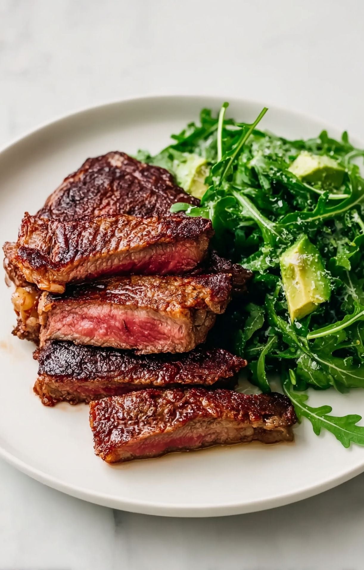 A clean white plate featuring a stack of sliced seared steak positioned next to a vibrant green arugula and avocado salad.