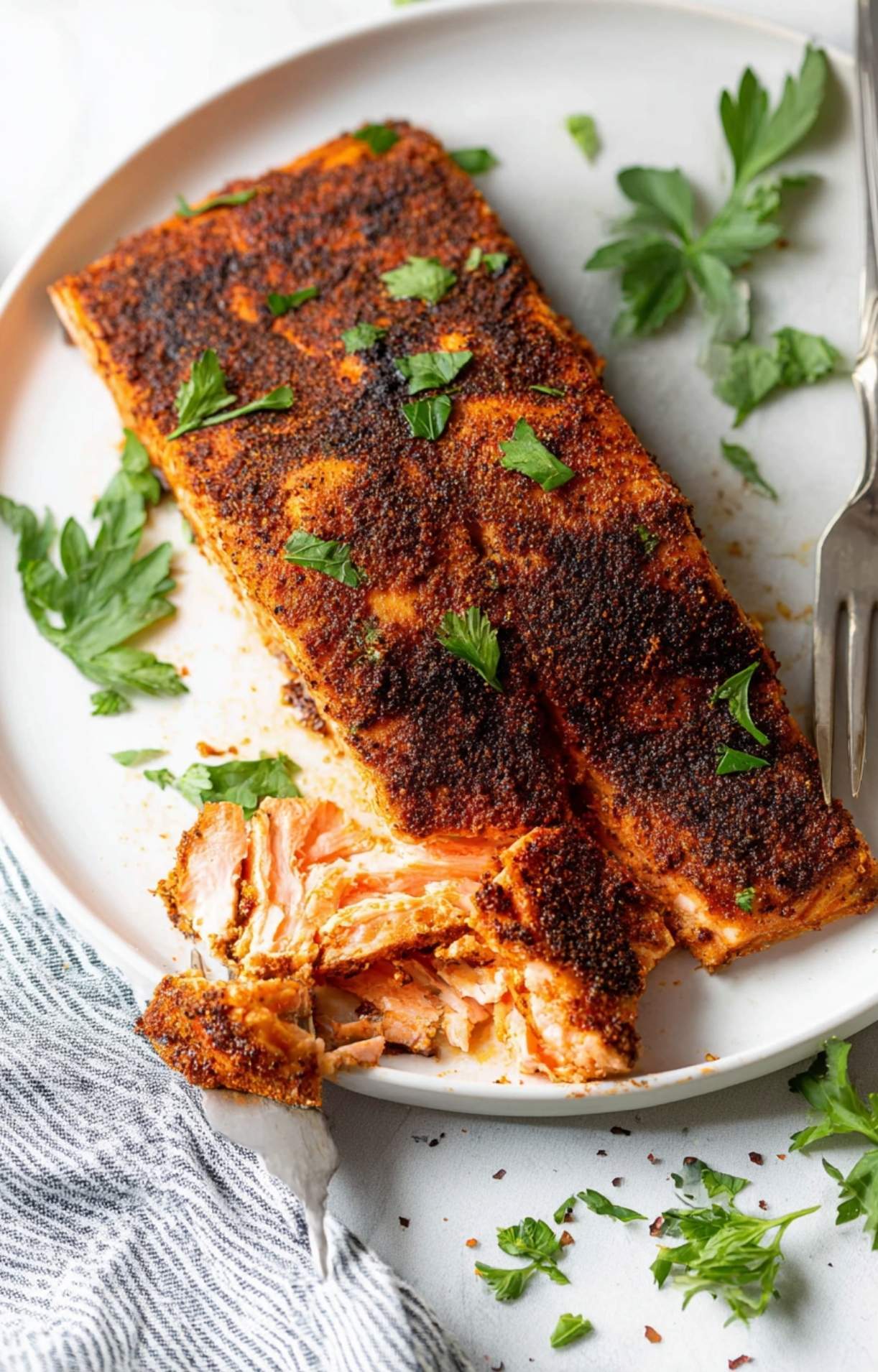 A cooked blackened salmon fillet on a white plate garnished with fresh parsley, with a fork pulling away a tender, flaky bite.