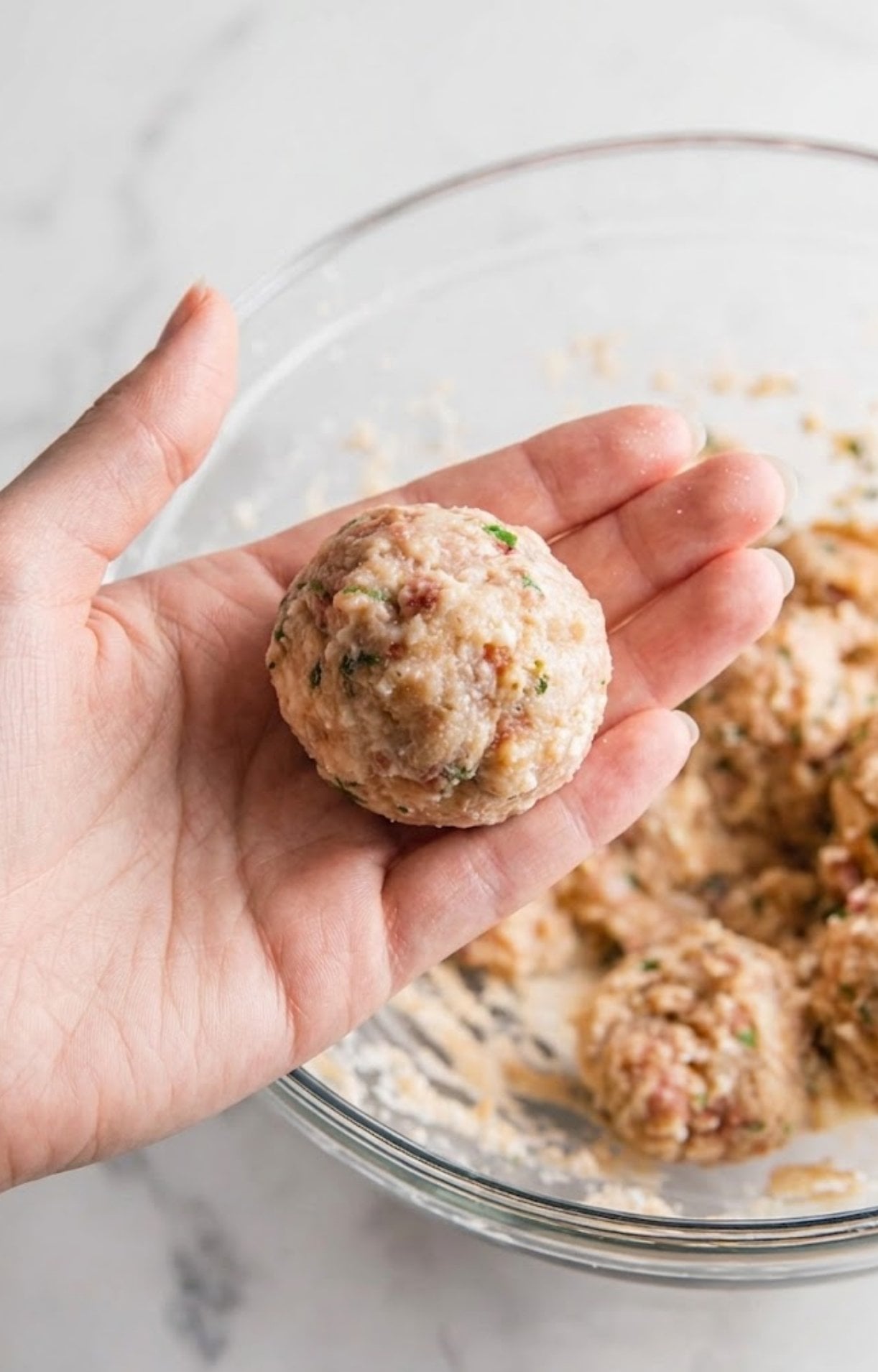 A hand holding a perfectly round, raw chicken meatball mixture flecked with green herbs over a glass mixing bowl.