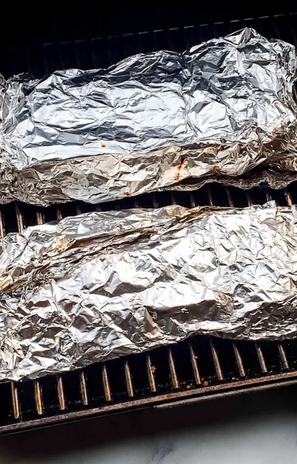 Two racks of ribs tightly wrapped in aluminum foil sitting on the pellet smoker grates to tenderize.