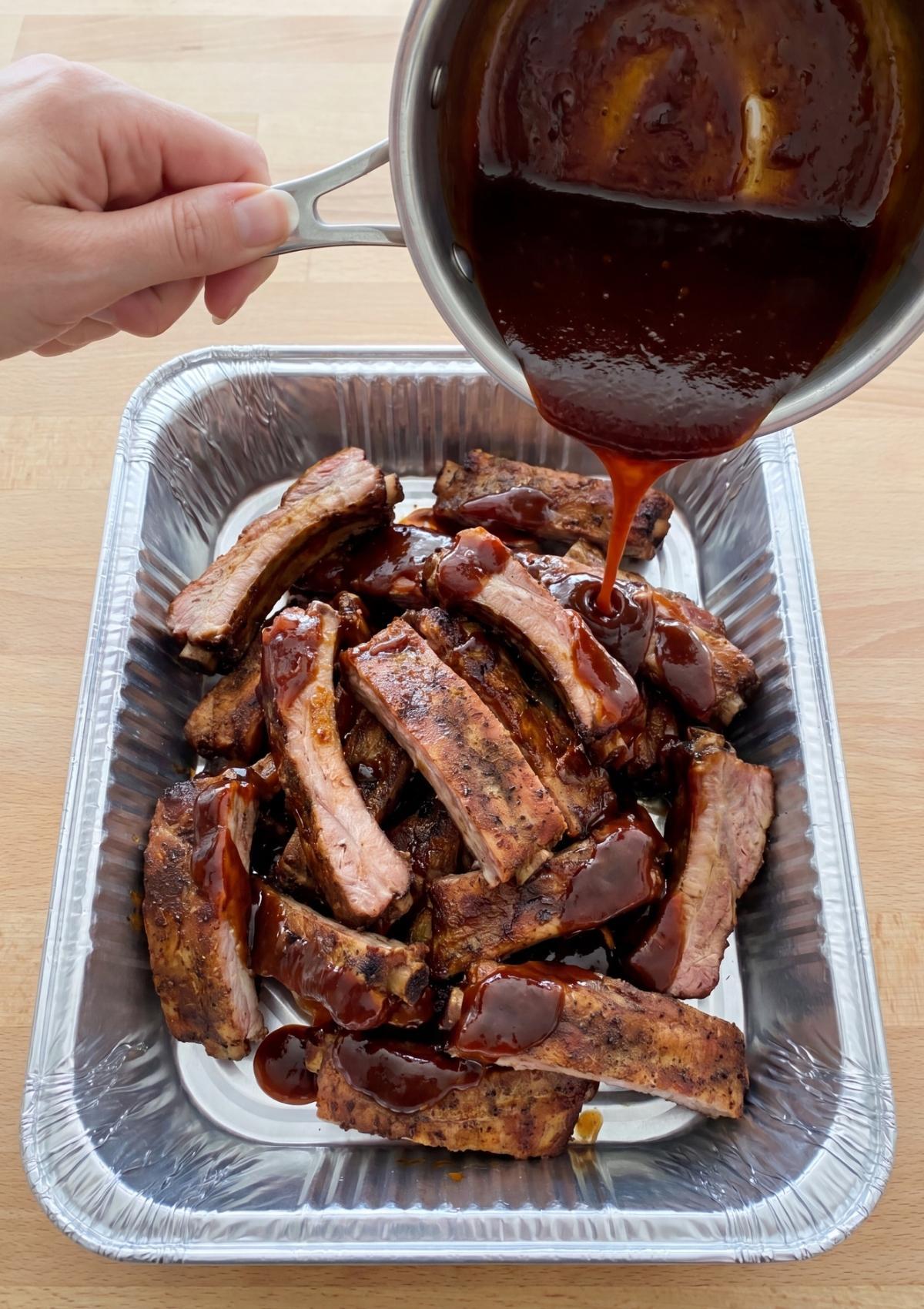 A hand pouring thick, dark barbecue sauce from a saucepan over a pile of smoked rib segments in a foil pan.