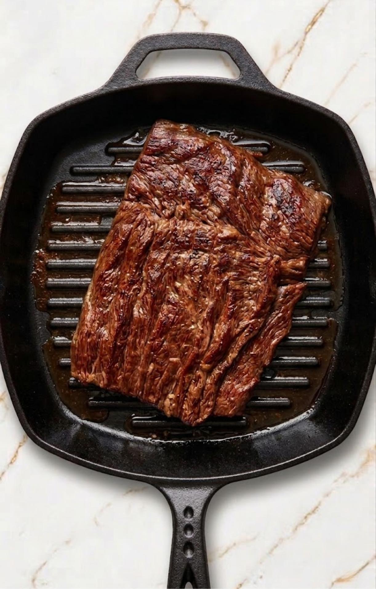 A seasoned flank steak cooking in a hot, cast-iron grill pan, showing browned edges and rendered juices.