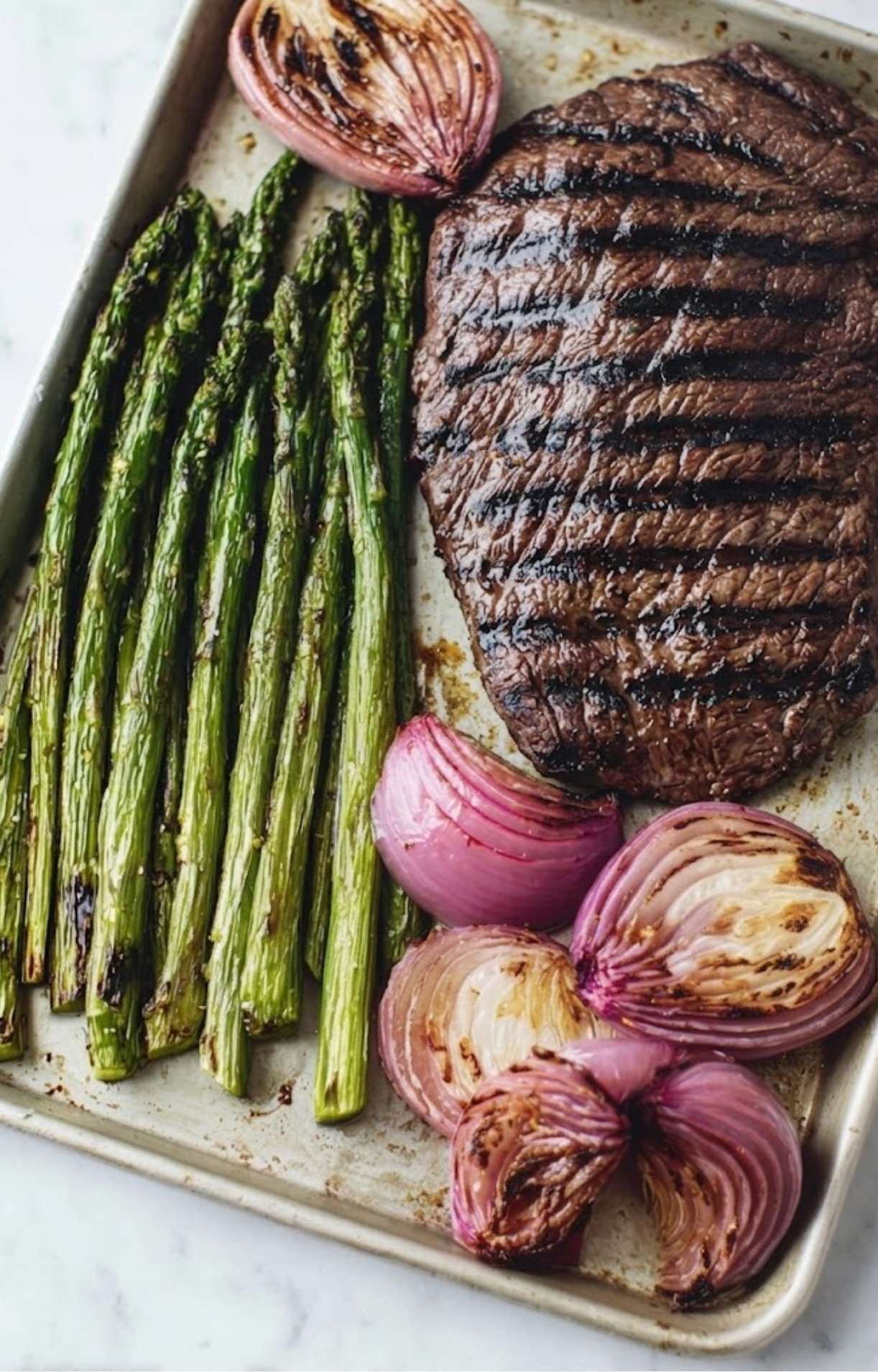 Grilled flank steak with charred sear marks resting on a tray next to roasted asparagus and softened red onion wedges.
