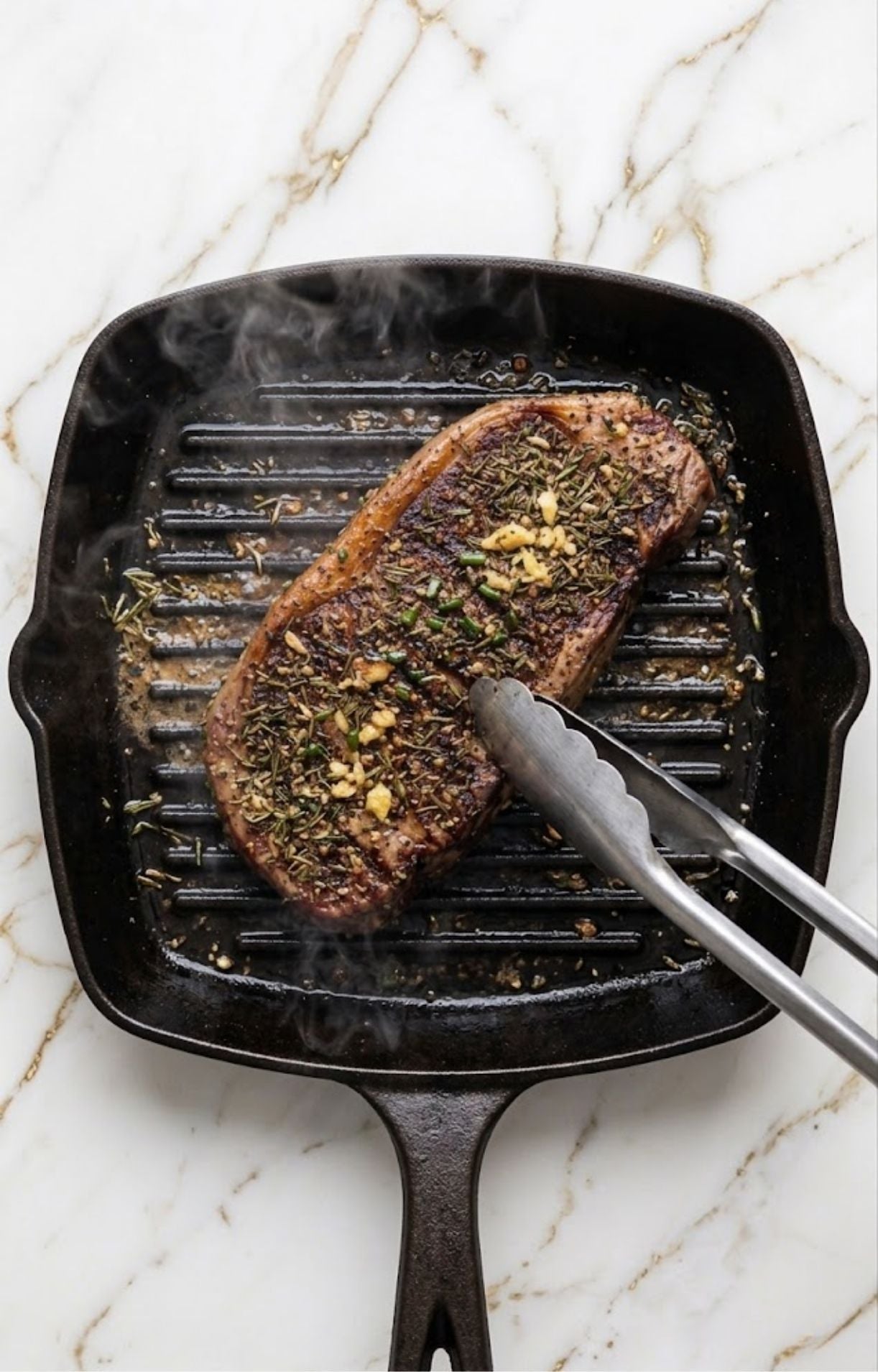 A seasoned steak being seared in a hot cast-iron grill pan with visible grill marks and steam, using metal tongs to flip the meat.
