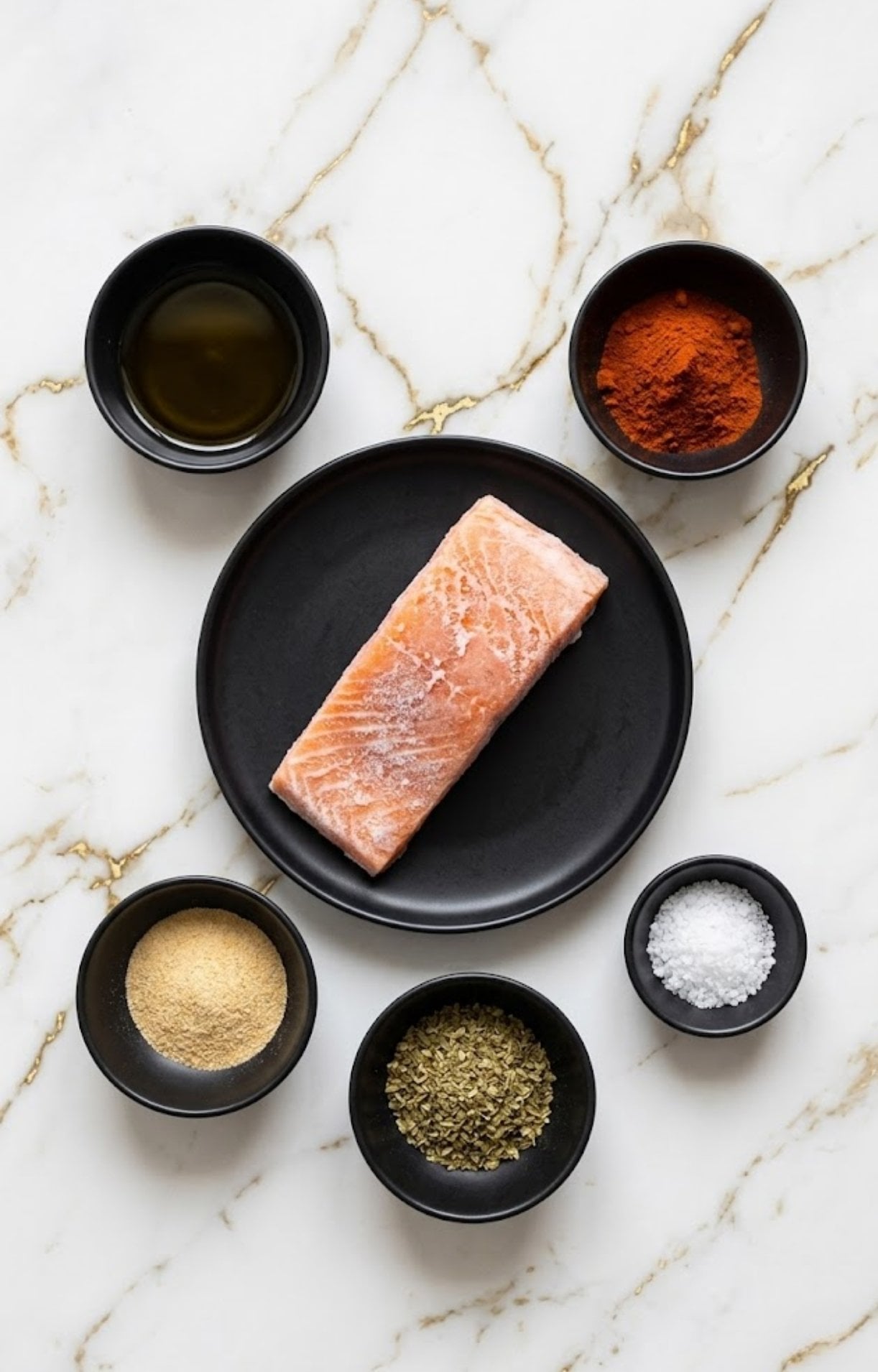 A flat lay of a frozen salmon fillet, olive oil, paprika, garlic powder, dried oregano, and sea salt in small black bowls on a marble surface.