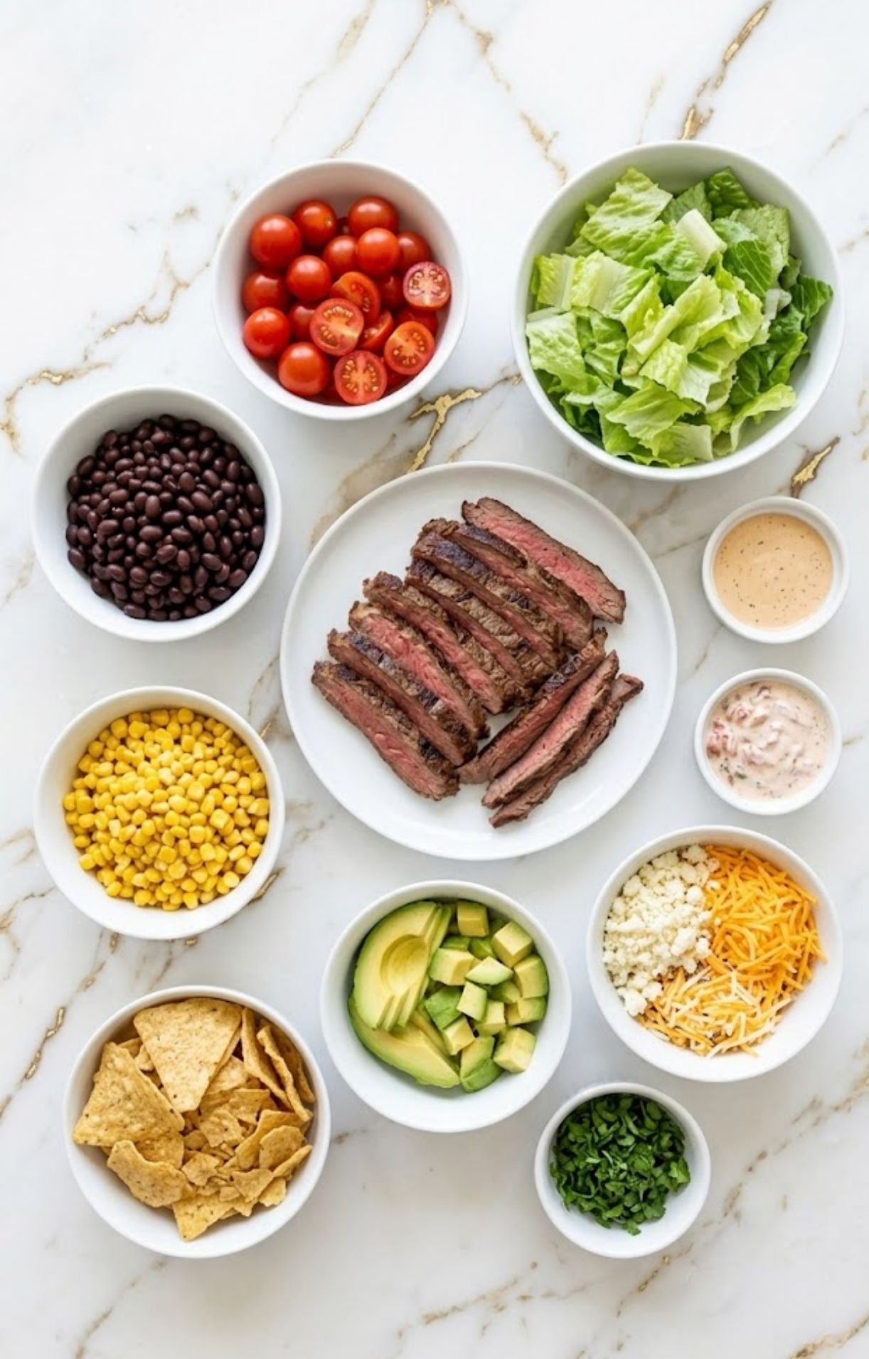 An overhead view of various bowls containing steak, corn, beans, tomatoes, lettuce, avocado, cheese, and dressing for an Easy Steak Taco Salad Recipe.