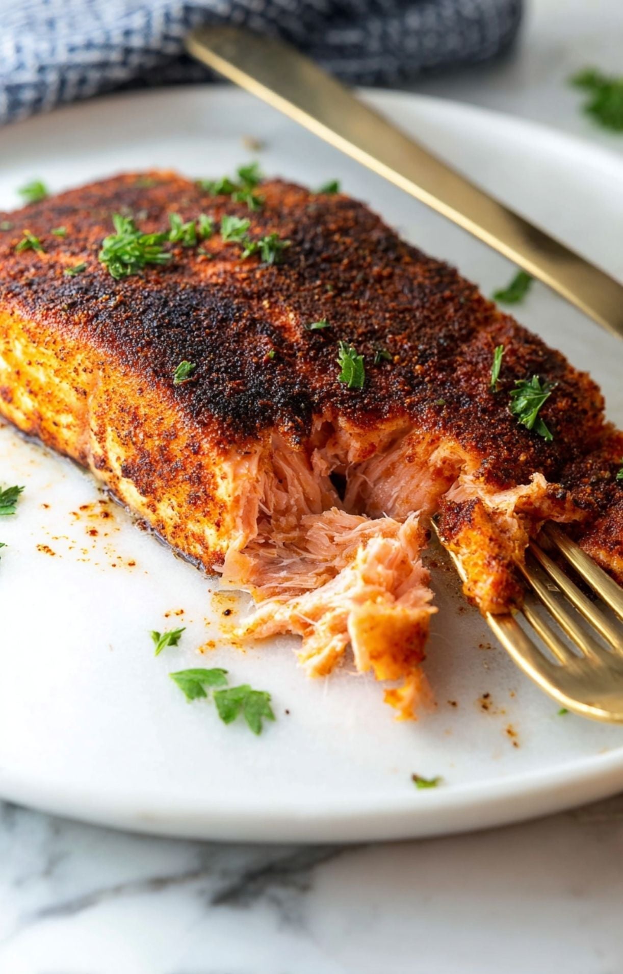 A beautiful close-up of a blackened salmon fillet showing the charred seasoning texture and juicy, pink interior being flaked by a gold fork.