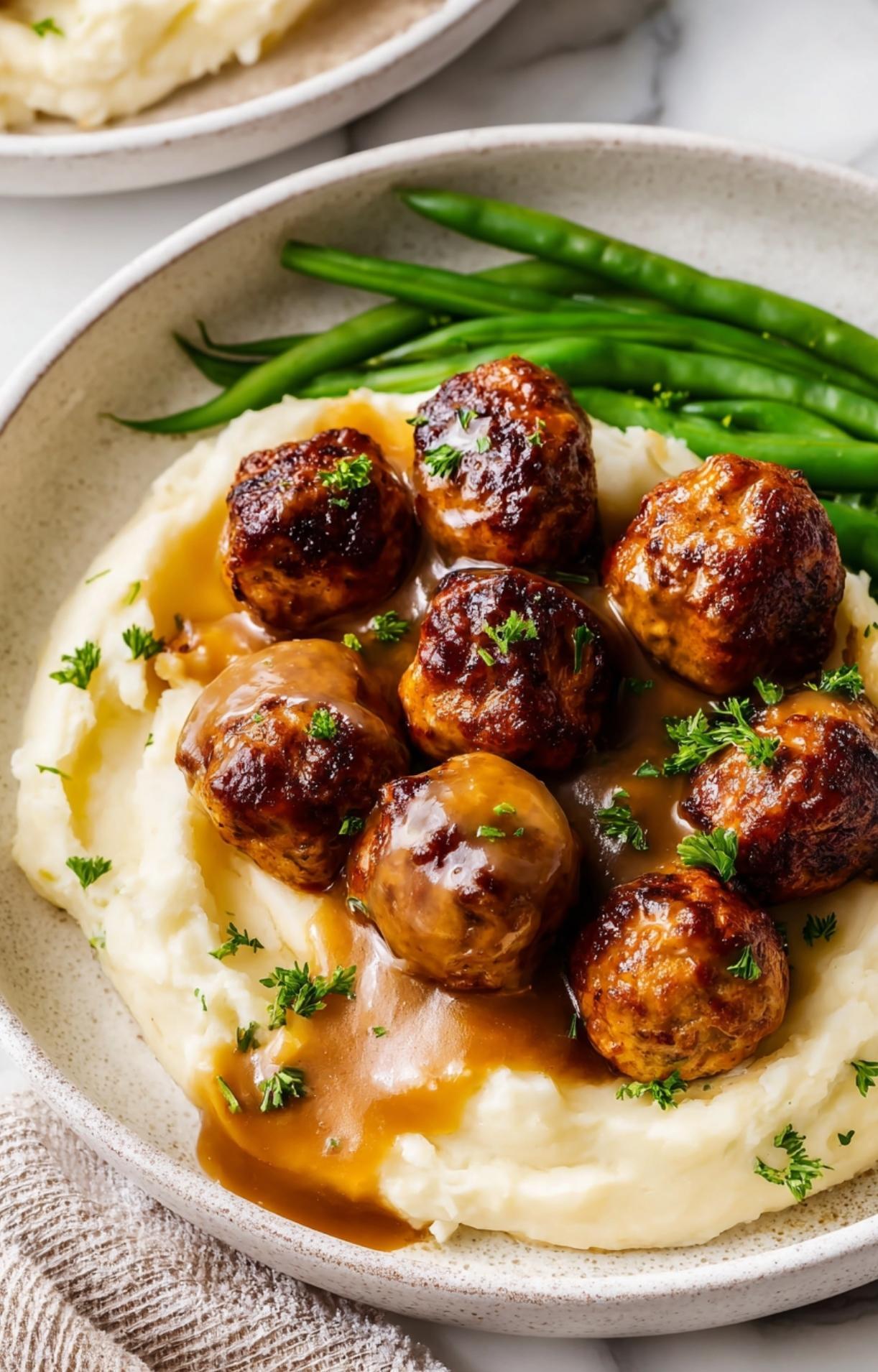 Close-up of herb-sprinkled chicken meatballs drizzled with brown gravy on a bed of fluffy mashed potatoes.