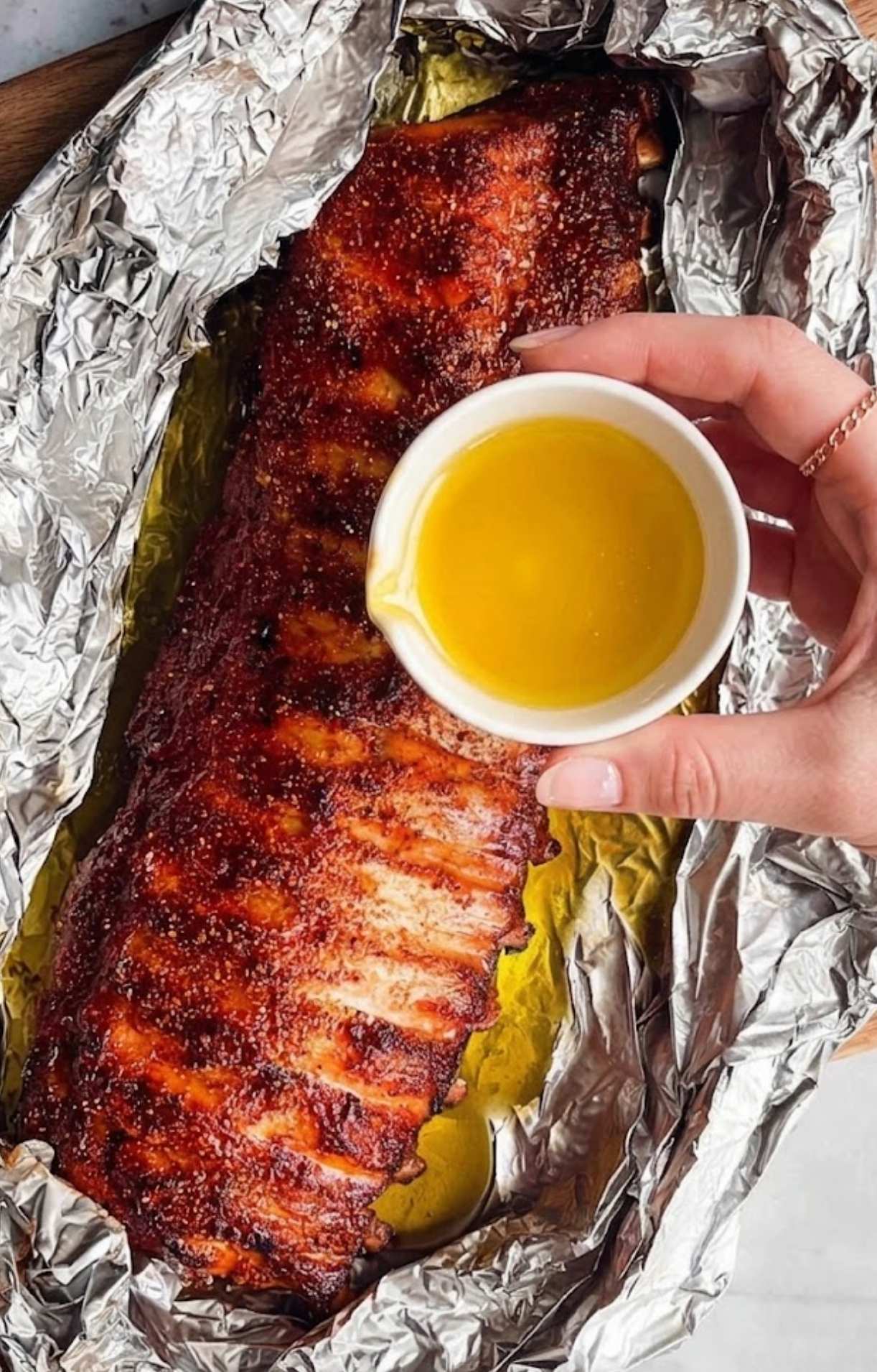 A hand with light pink nails pouring liquid over a rack of baby back ribs inside an open foil packet.