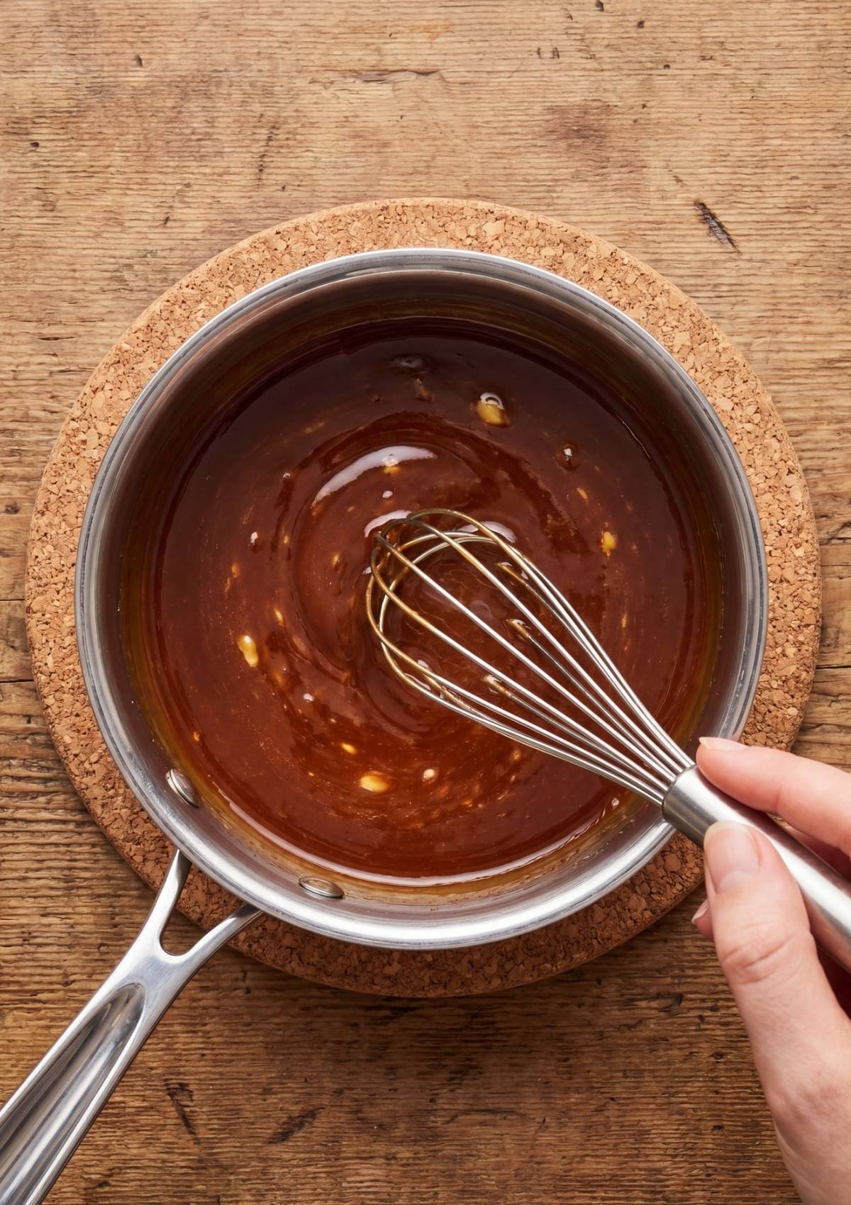 A hand whisking a glossy, mahogany-colored BBQ sauce in a stainless steel pot on a wooden surface.