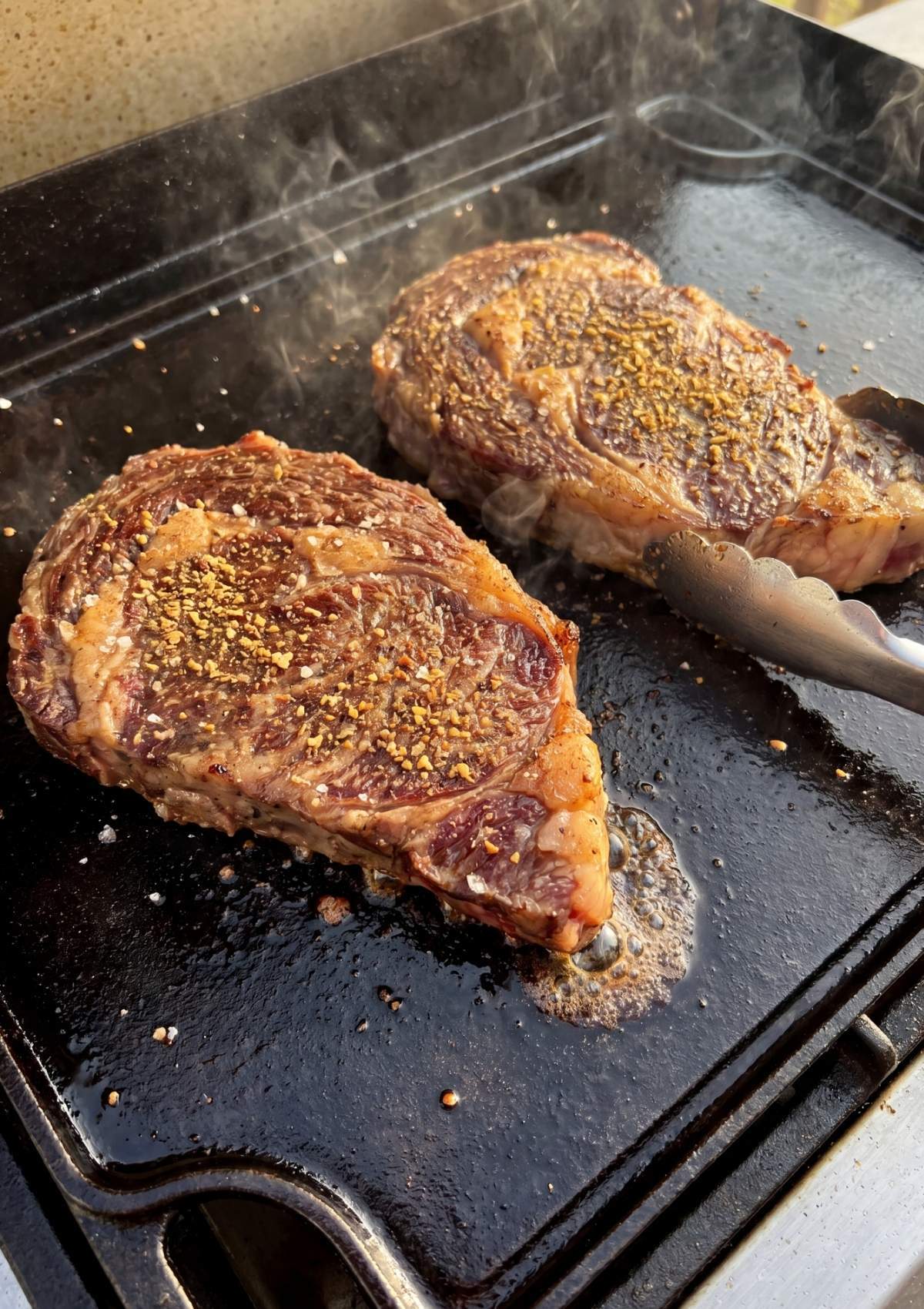 Two Wagyu steaks searing on a hot flat-top griddle with visible steam, herbs, and rendered fat bubbling around the edges.