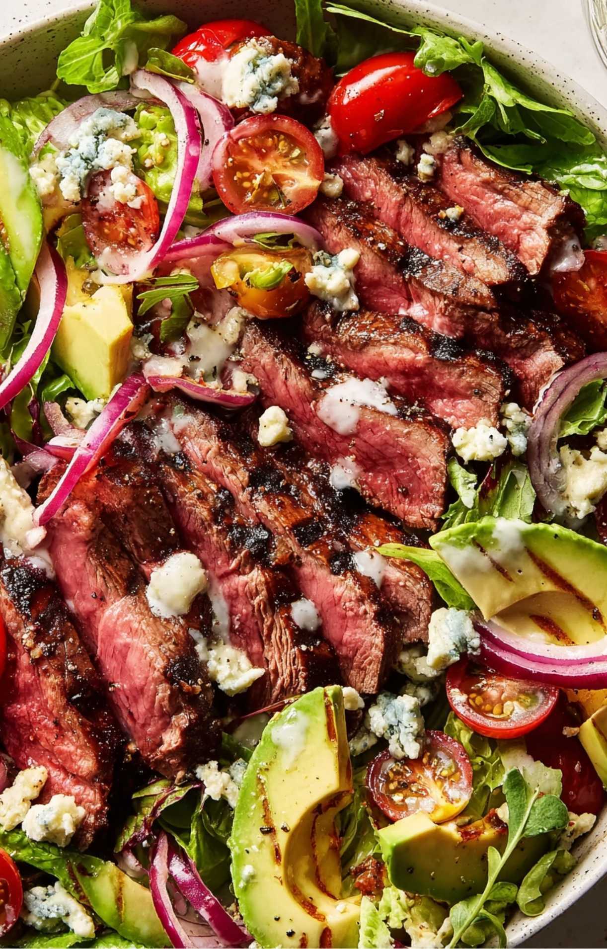 A vibrant bowl showing sliced steak layered over greens with tomatoes, avocado, red onion, and fresh blue cheese crumbles.