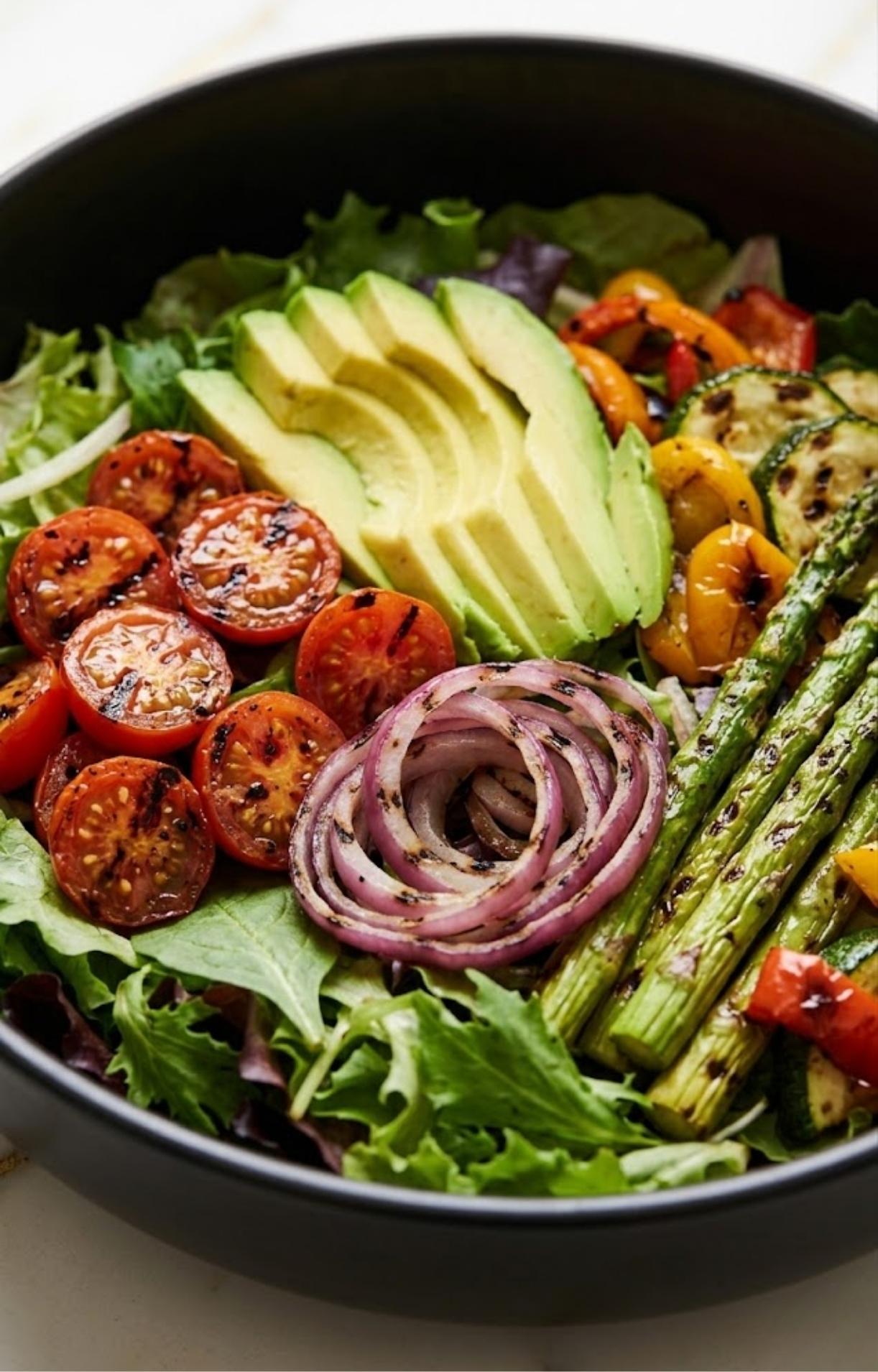 A large black bowl filled with fresh greens topped with grilled tomatoes, asparagus, zucchini, red onions, and sliced avocado.