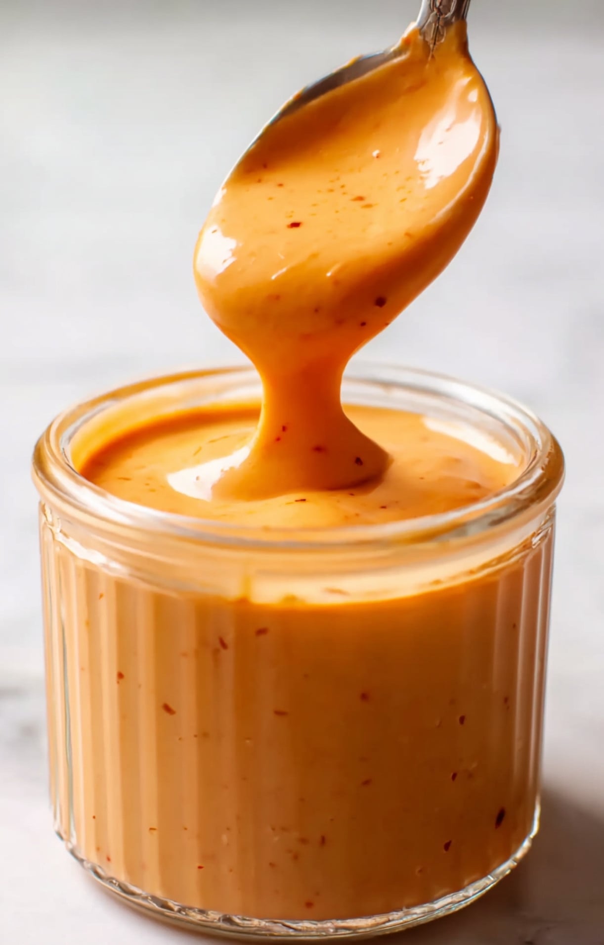 A close-up of a spoon lifting creamy orange sauce out of a glass jar.