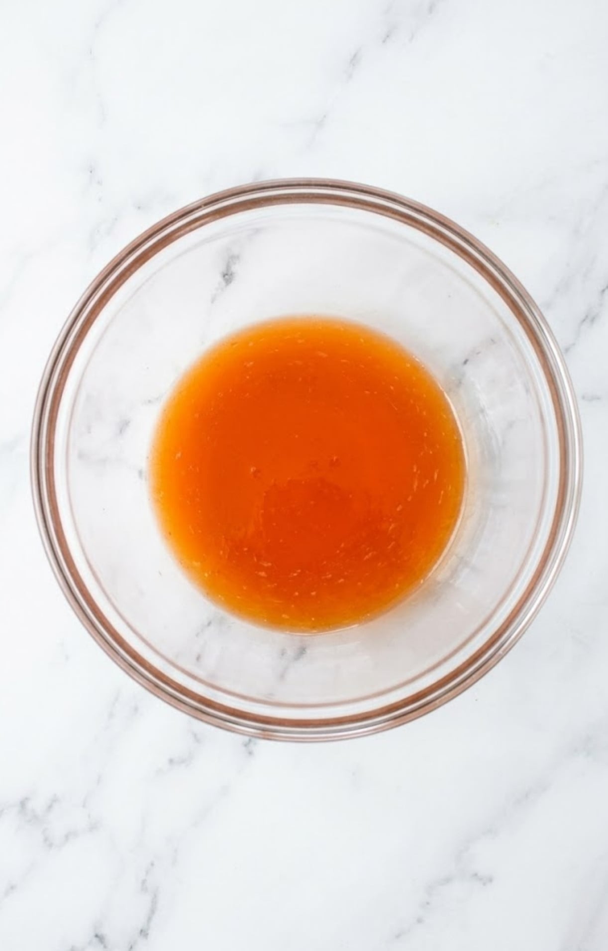 A clear glass mixing bowl filled with an orange-tinted liquid sauce ready to coat frozen meatballs air fryer.