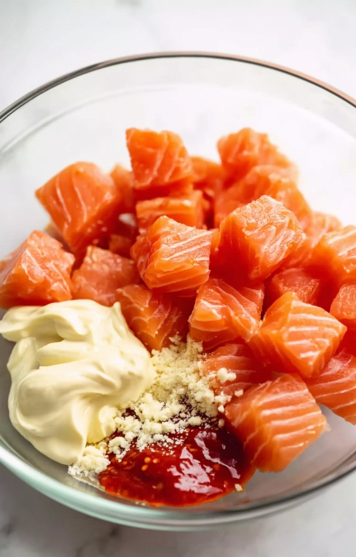 Raw cubes of fresh salmon in a glass bowl being tossed with mayonnaise, sriracha, and seasoning for an Air Fryer Salmon Bowl.