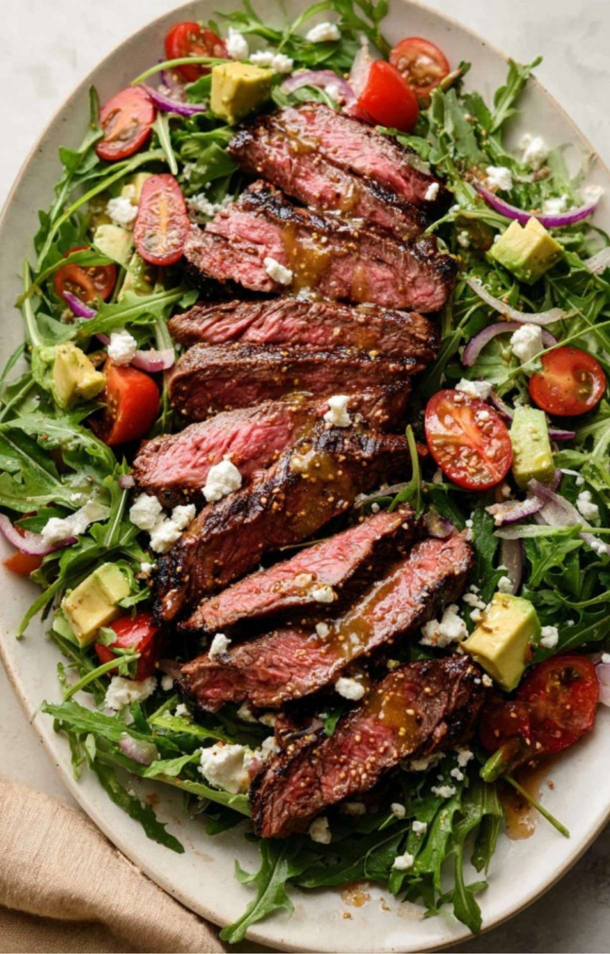 An overhead view of a large platter featuring seasoned steak strips laid over greens with red onions, tomatoes, and feta.