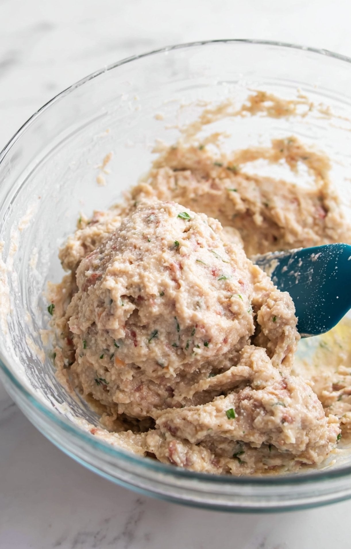Ground chicken mixture being combined with herbs and binders in a glass bowl using a blue spatula.