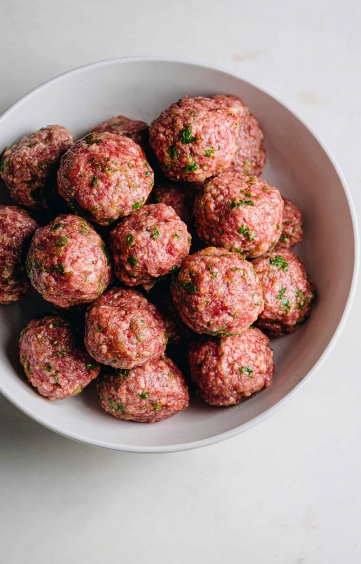 A white bowl filled with hand-rolled raw lamb kofta meatballs showing visible flecks of fresh green herbs.