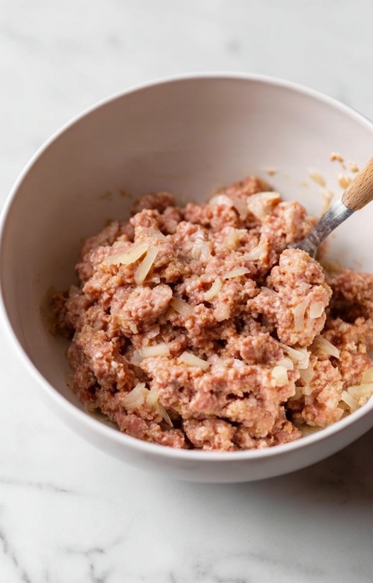 A light pink mixing bowl filled with ground pork meat and diced onions being combined with a wooden-handled spoon.
