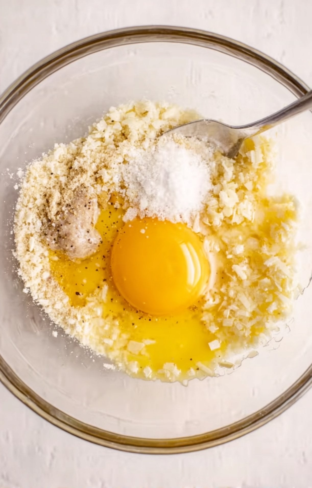 A glass bowl containing an egg, breadcrumbs, minced garlic, salt, and pepper being mixed with a spoon.