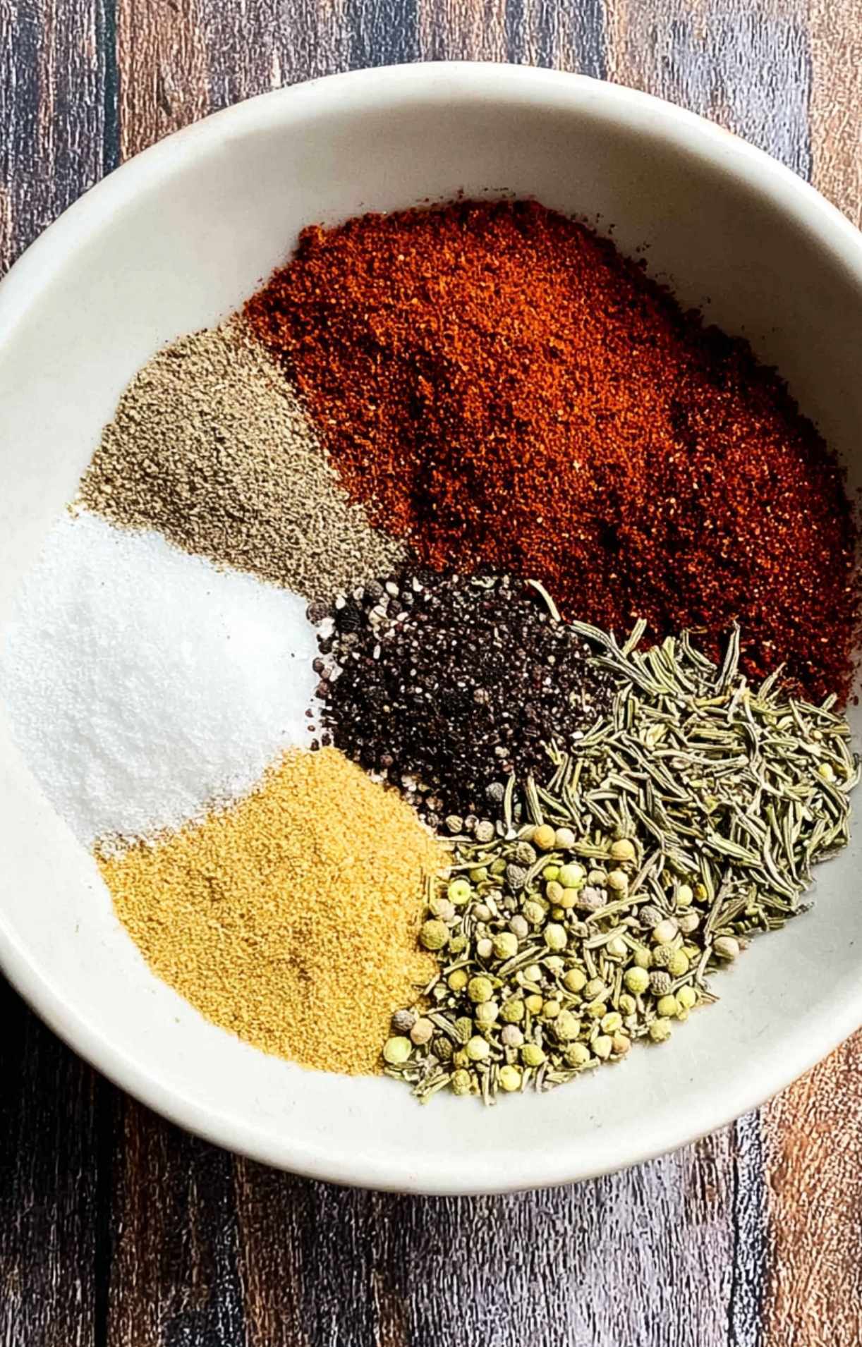 A white bowl containing distinct piles of spices, including paprika, garlic powder, salt, black pepper, and dried rosemary.