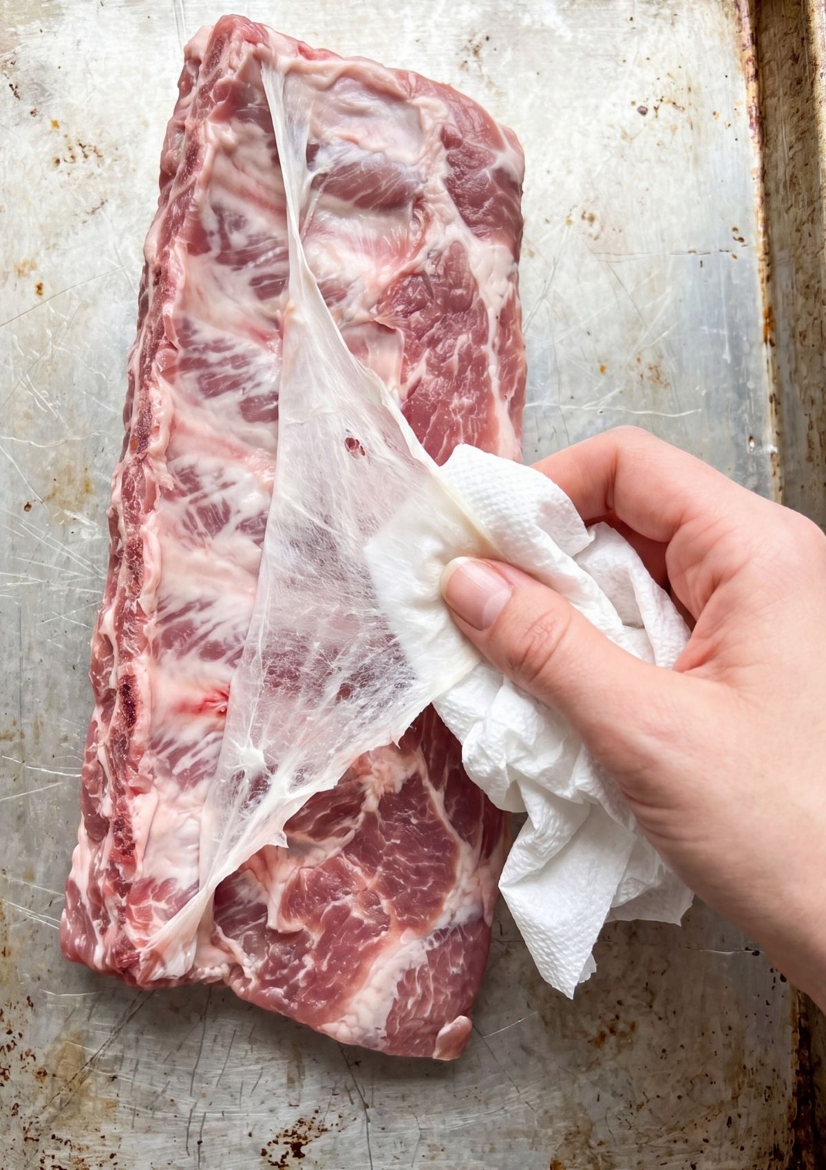 A hand using a paper towel to peel the silver skin membrane off a raw rack of pork ribs on a baking sheet.