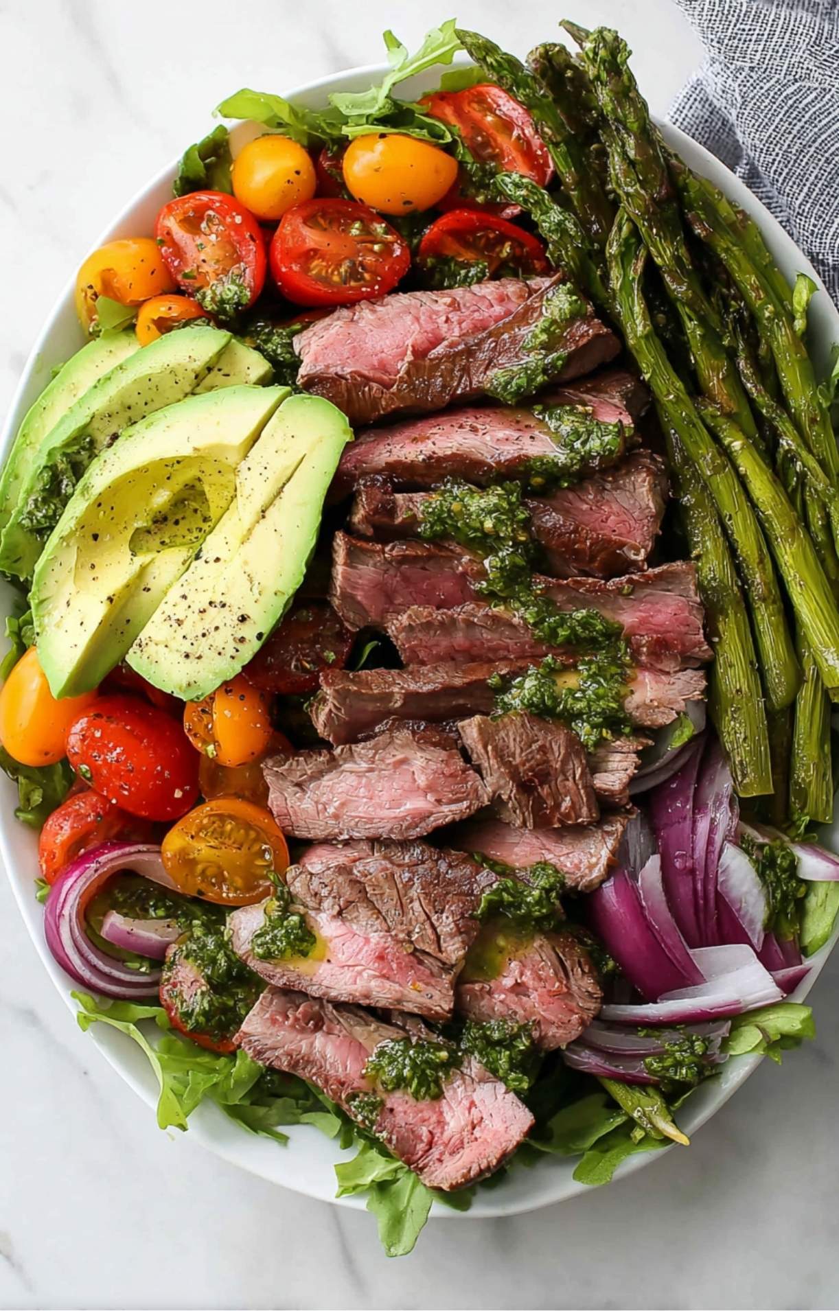 A close-up of a white platter featuring sliced medium-rare steak drizzled with green chimichurri sauce over a bed of salad.