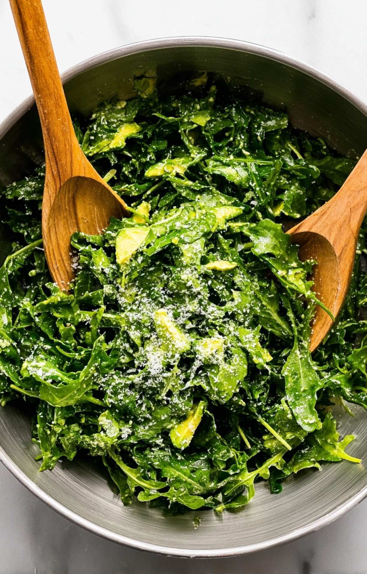 Two wooden salad servers mixing the arugula, avocado, and Parmesan cheese with a lemon-garlic dressing.