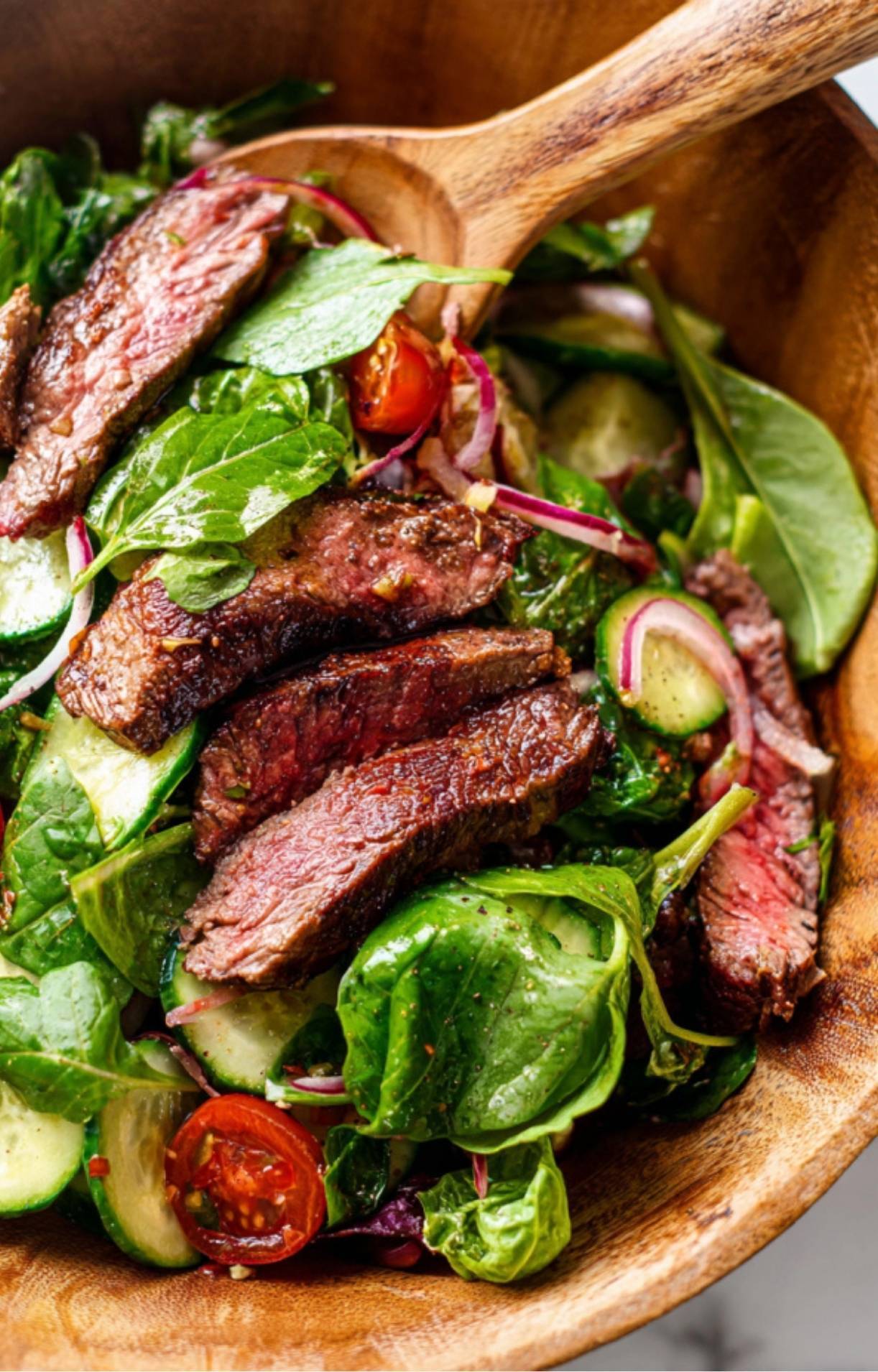 A wooden spoon tossing fresh spinach, arugula, and steak slices together in a large bowl to distribute the Steak Salad Recipe dressing.