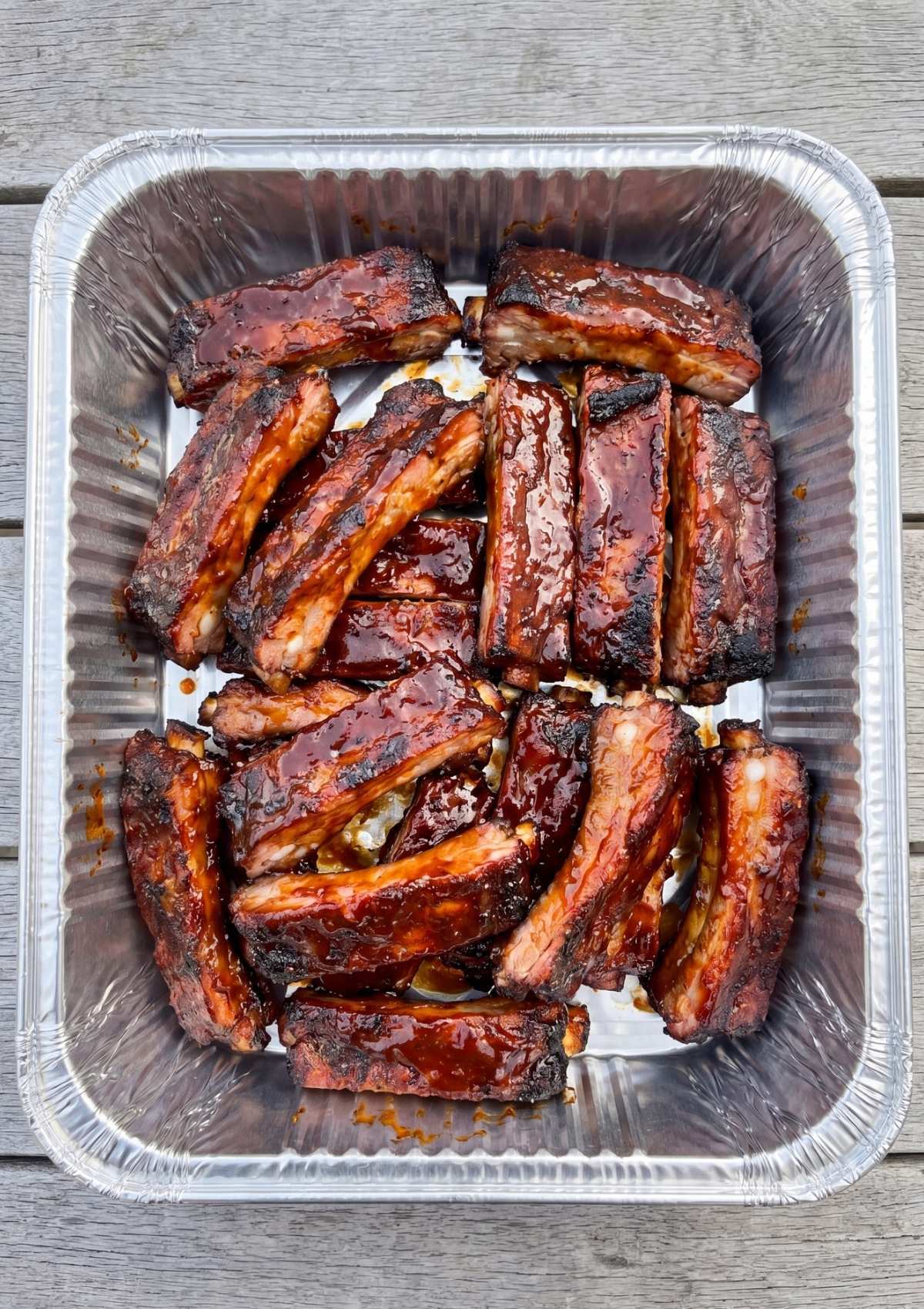 A silver disposable foil pan filled with sticky, barbecue-glazed individual rib segments sitting on a rustic grey wooden outdoor table.