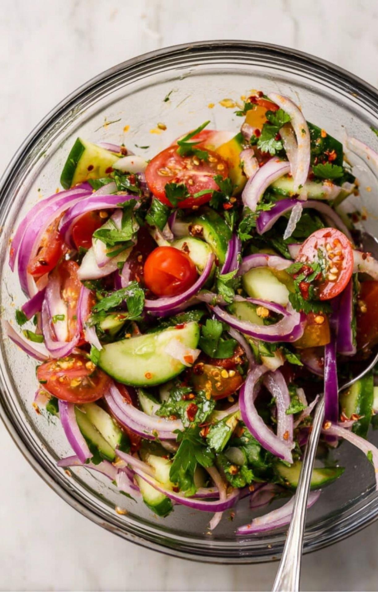 A glass mixing bowl filled with sliced red onions, cucumbers, cherry tomatoes, and fresh cilantro tossed in a light dressing.