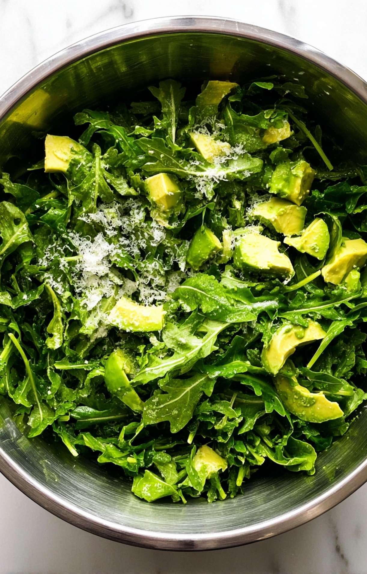 A stainless steel mixing bowl filled with fresh baby arugula leaves topped with chunks of ripe avocado and grated cheese.