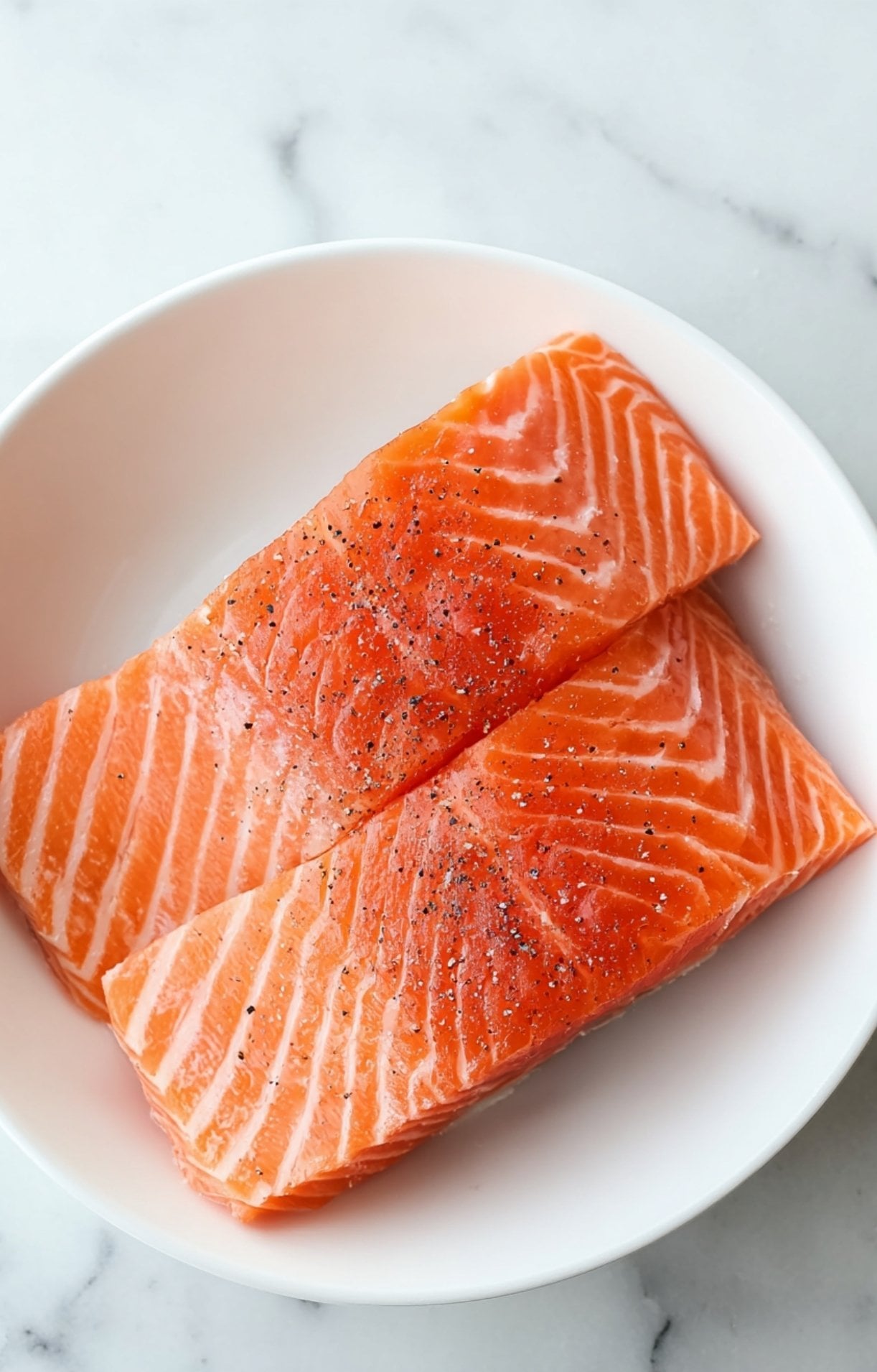 Two fresh, raw salmon fillets seasoned with salt and black pepper in a white bowl before air frying.