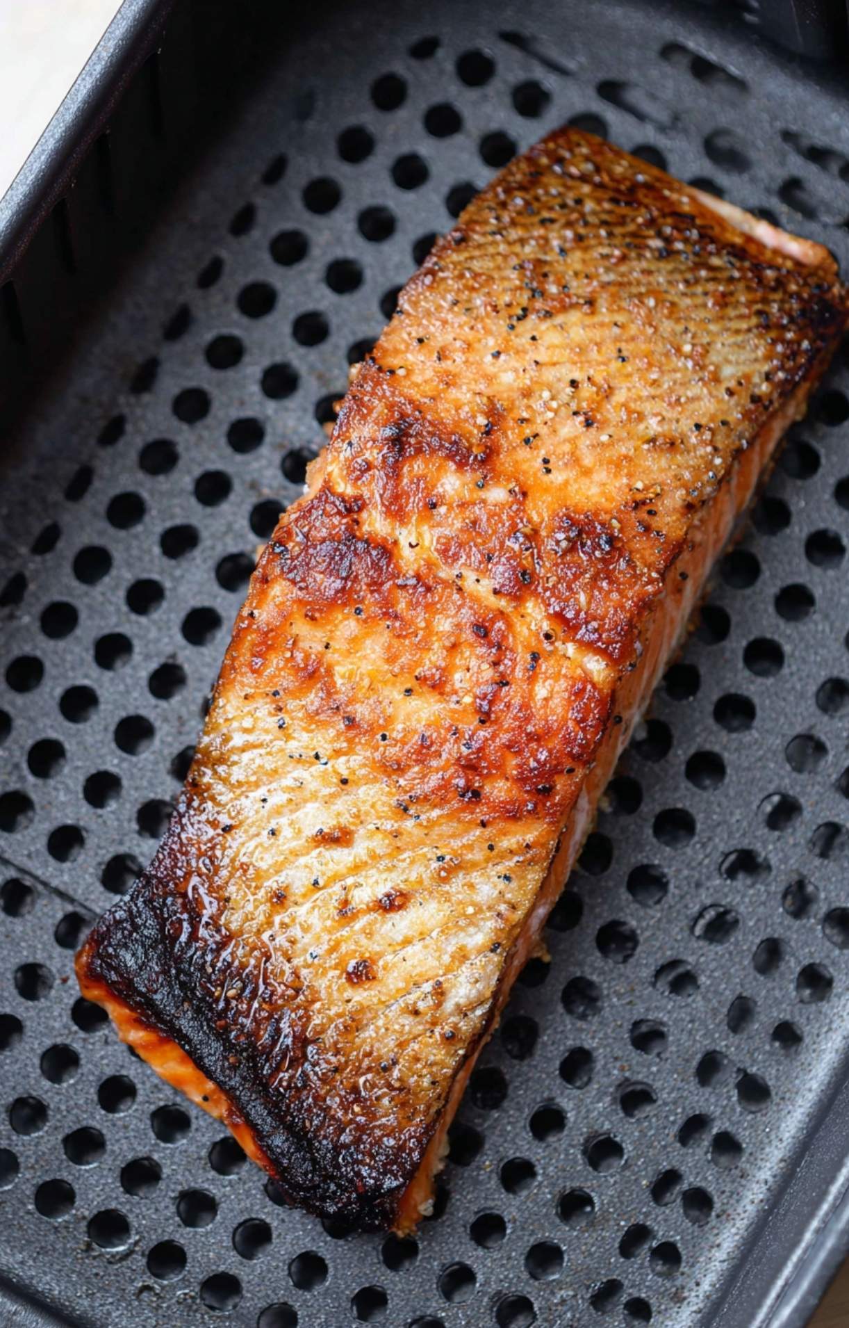 A single salmon fillet cooking inside a black air fryer basket, showing the skin beginning to crisp and brown.