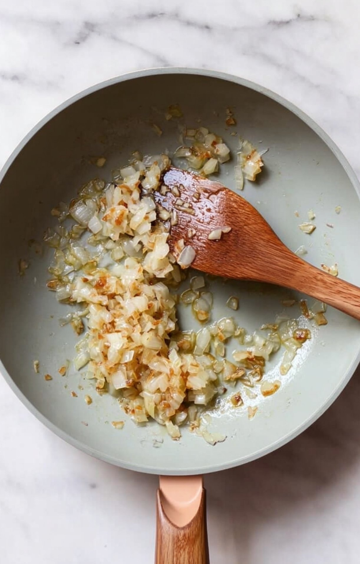 Sautéed diced onions being stirred with a wooden spatula in a grey non-stick skillet on a marble surface.