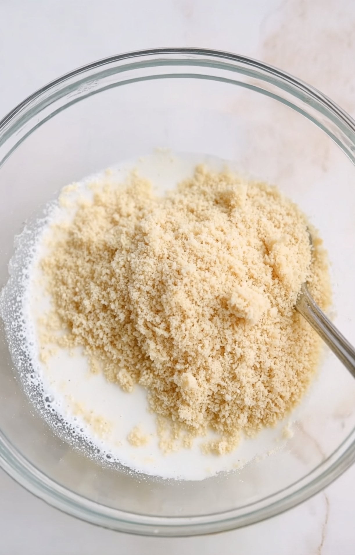 A glass mixing bowl showing white milk being poured over breadcrumbs to create a panade for moist meatballs.