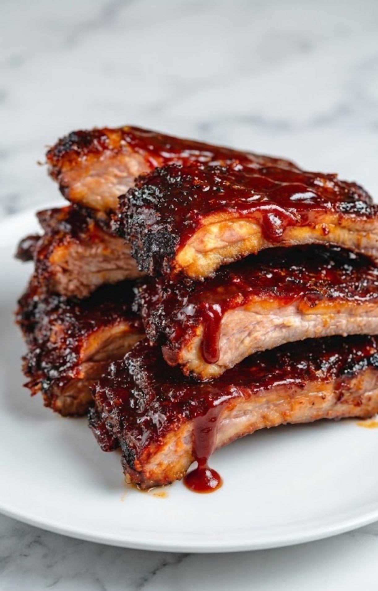 Close-up of tender baby back ribs with BBQ sauce dripping down on a white plate and marble surface.