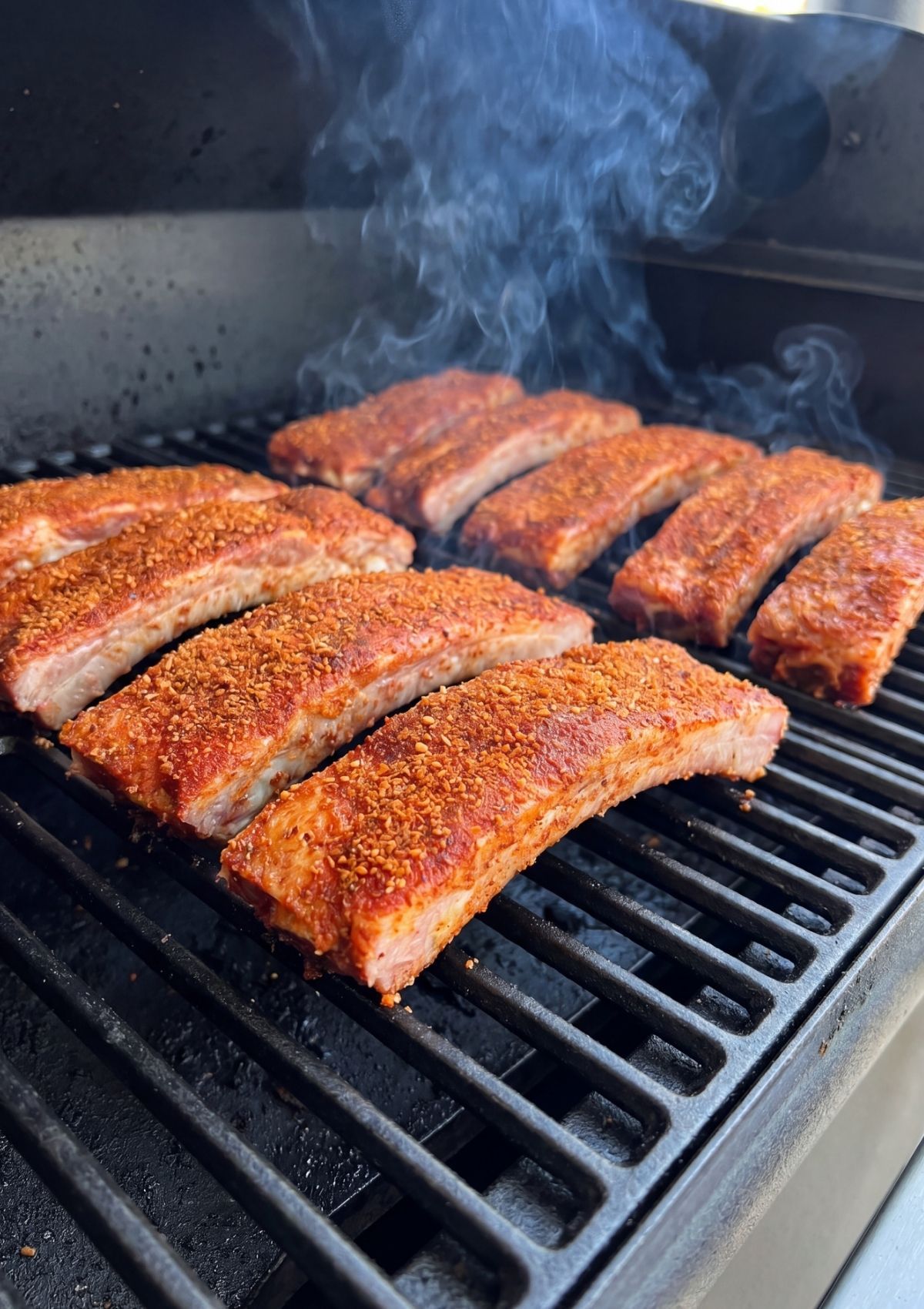 Row of individual seasoned ribs smoking on the black grates of a pellet grill with visible wisps of blue smoke.