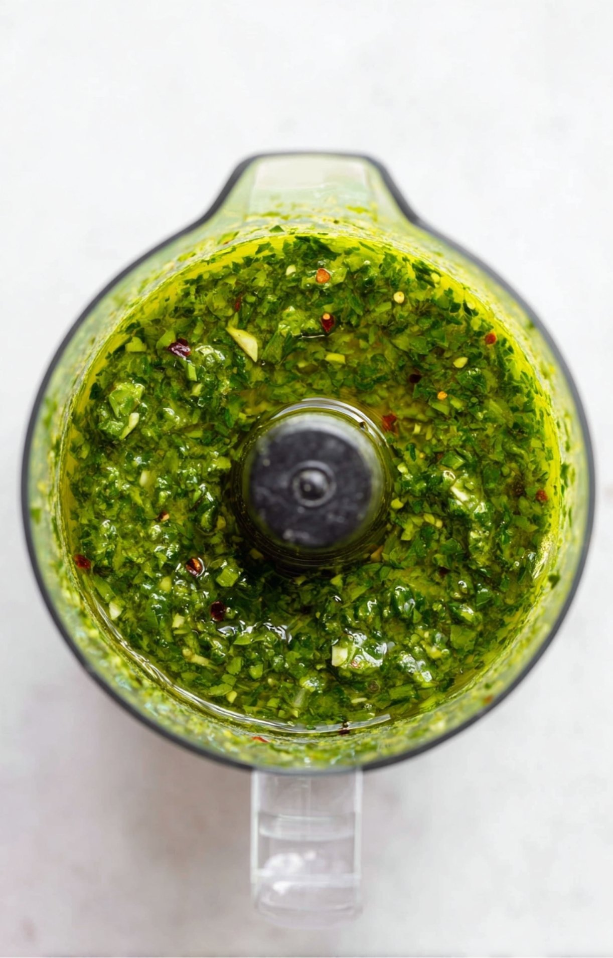 An overhead view of a food processor filled with a bright green, herbaceous chimichurri sauce made of parsley, garlic, and oil.