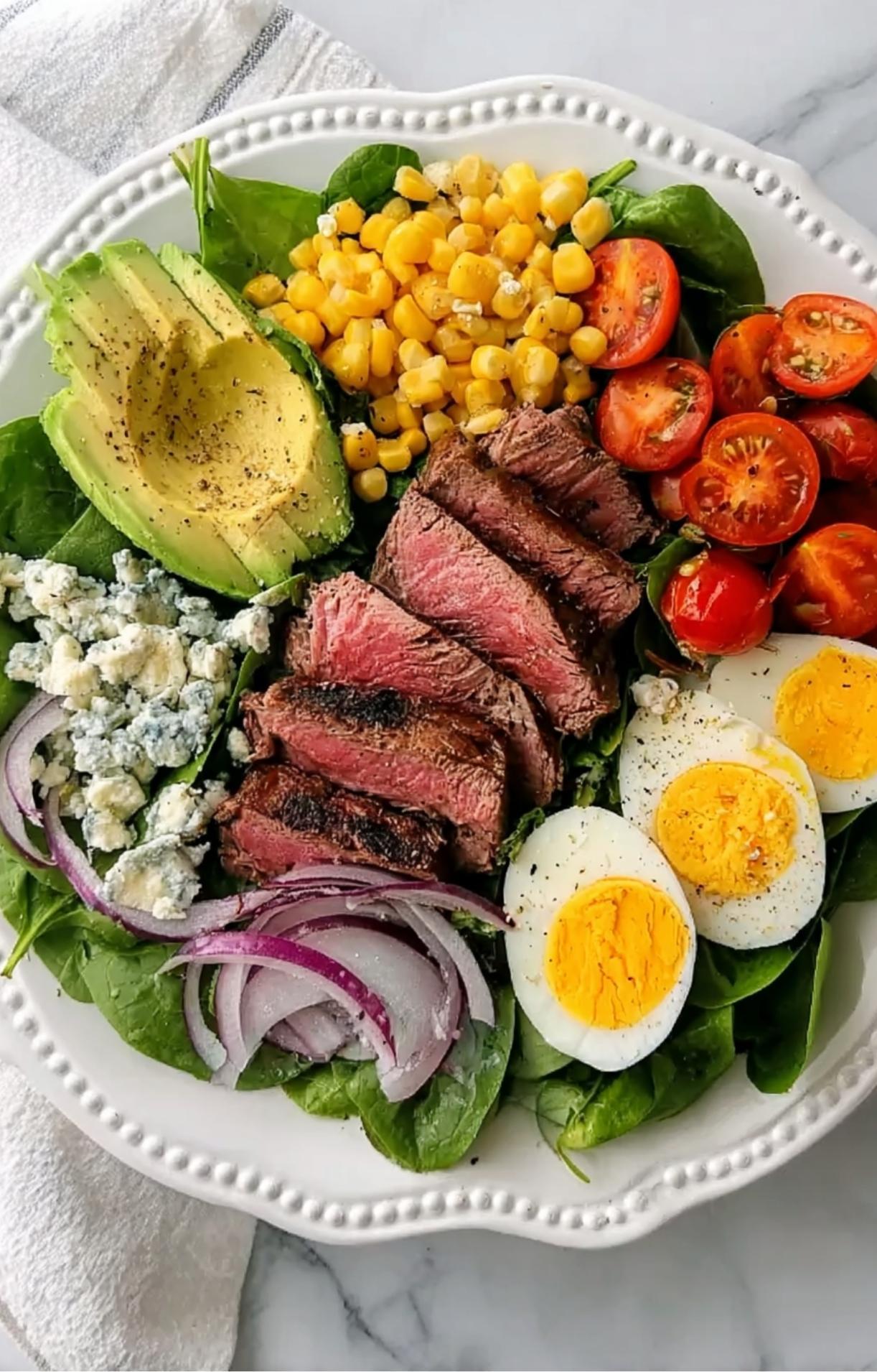 A white bowl filled with a bed of spinach topped with sliced steak, boiled eggs, cherry tomatoes, grilled corn, avocado, blue cheese, and red onions.
