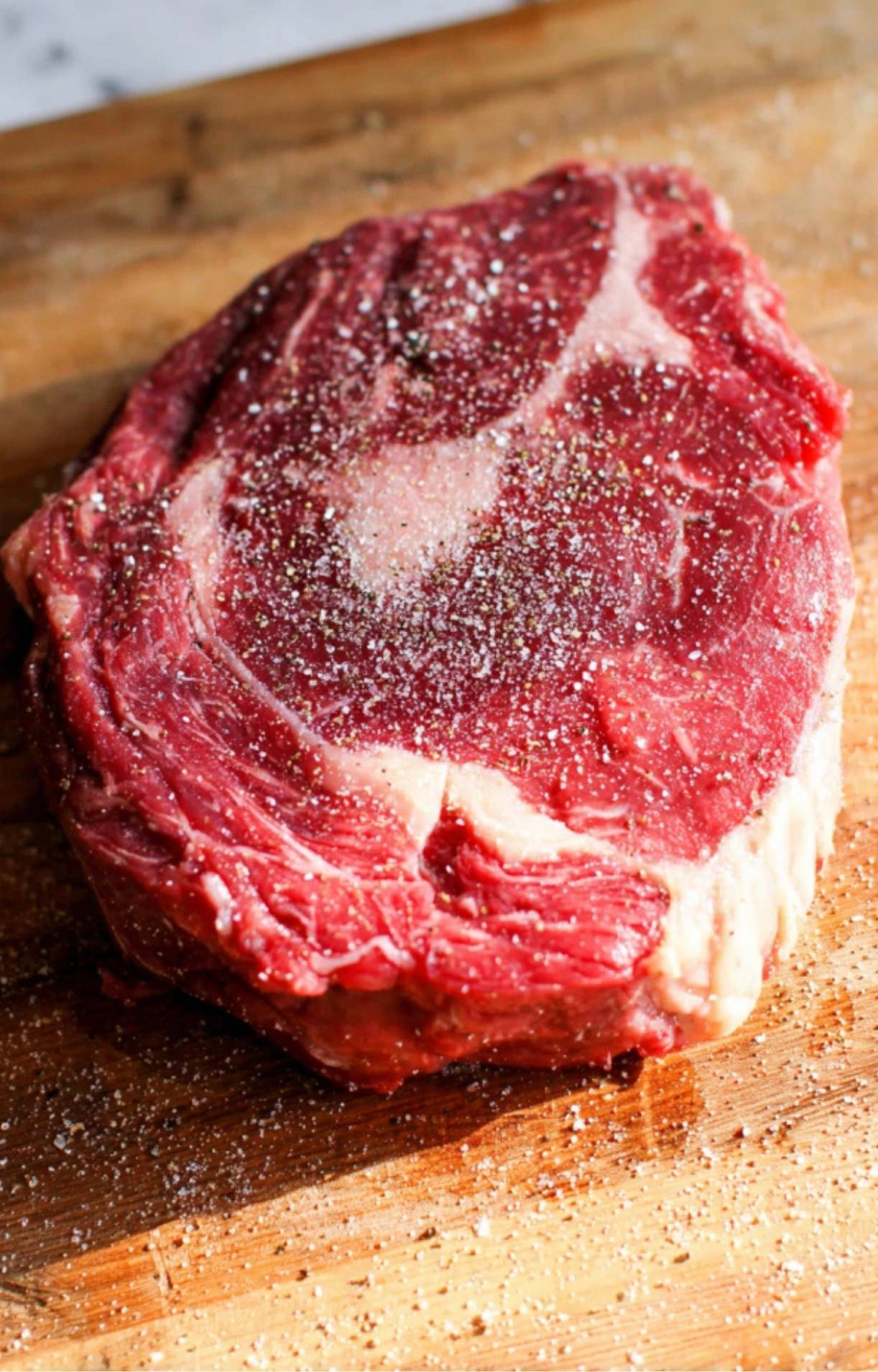 A raw ribeye steak placed on a wooden cutting board and generously seasoned with coarse salt and cracked black pepper for a Steak Salad Recipe.
