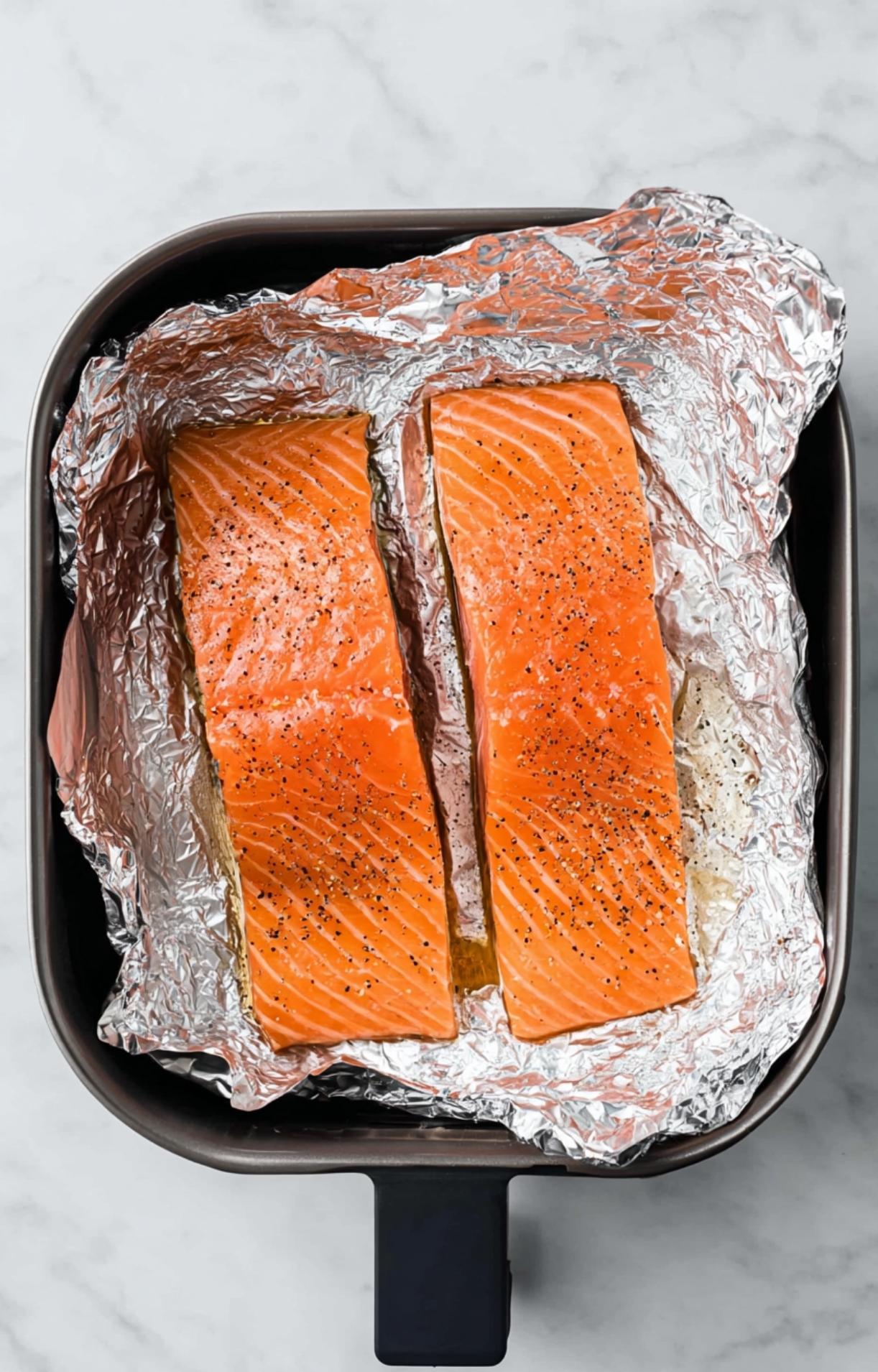 Two seasoned salmon fillets placed on aluminum foil inside an air fryer basket, ready to begin the cooking process.