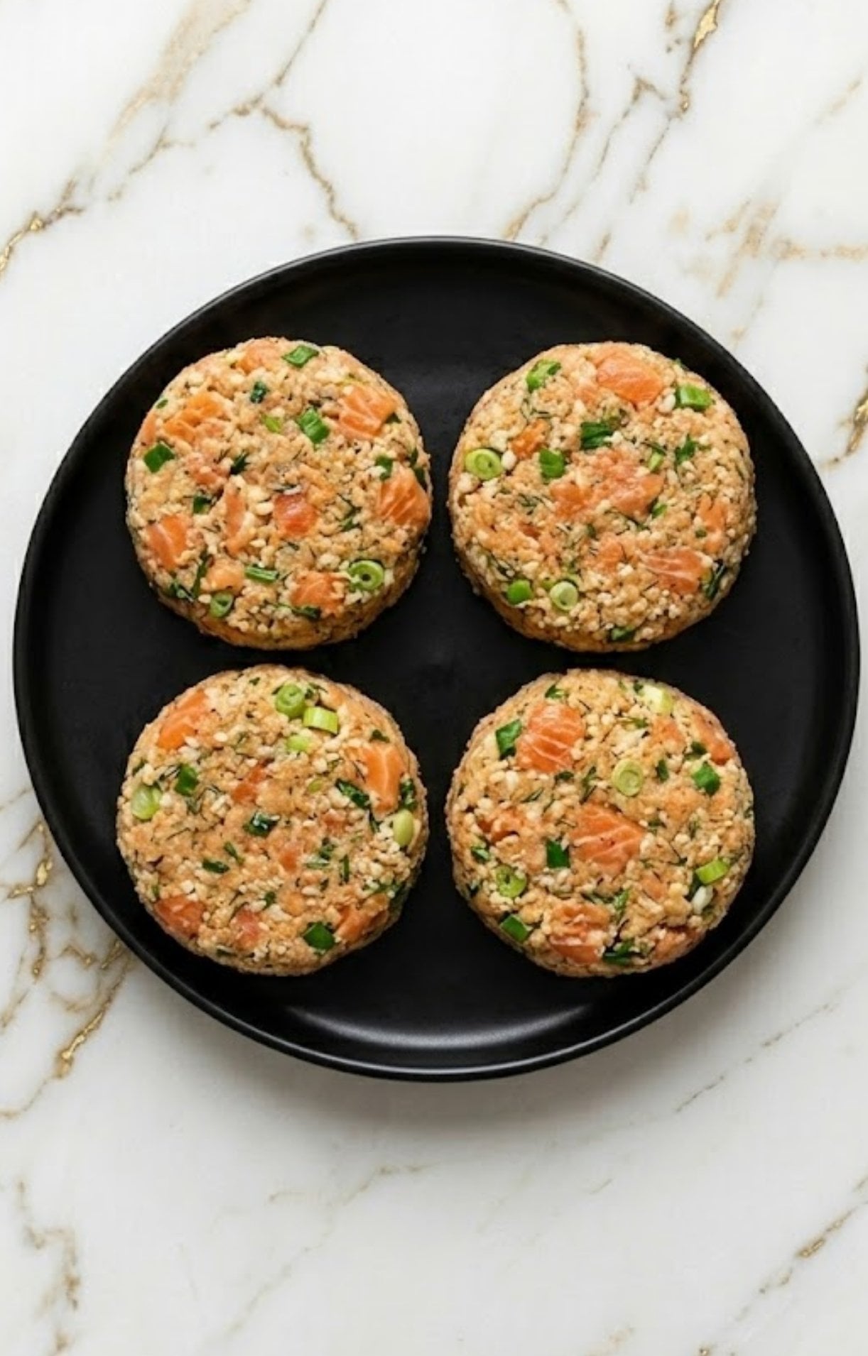 Four raw salmon patties mixed with green onions and herbs, formed into discs and placed on a black plate.