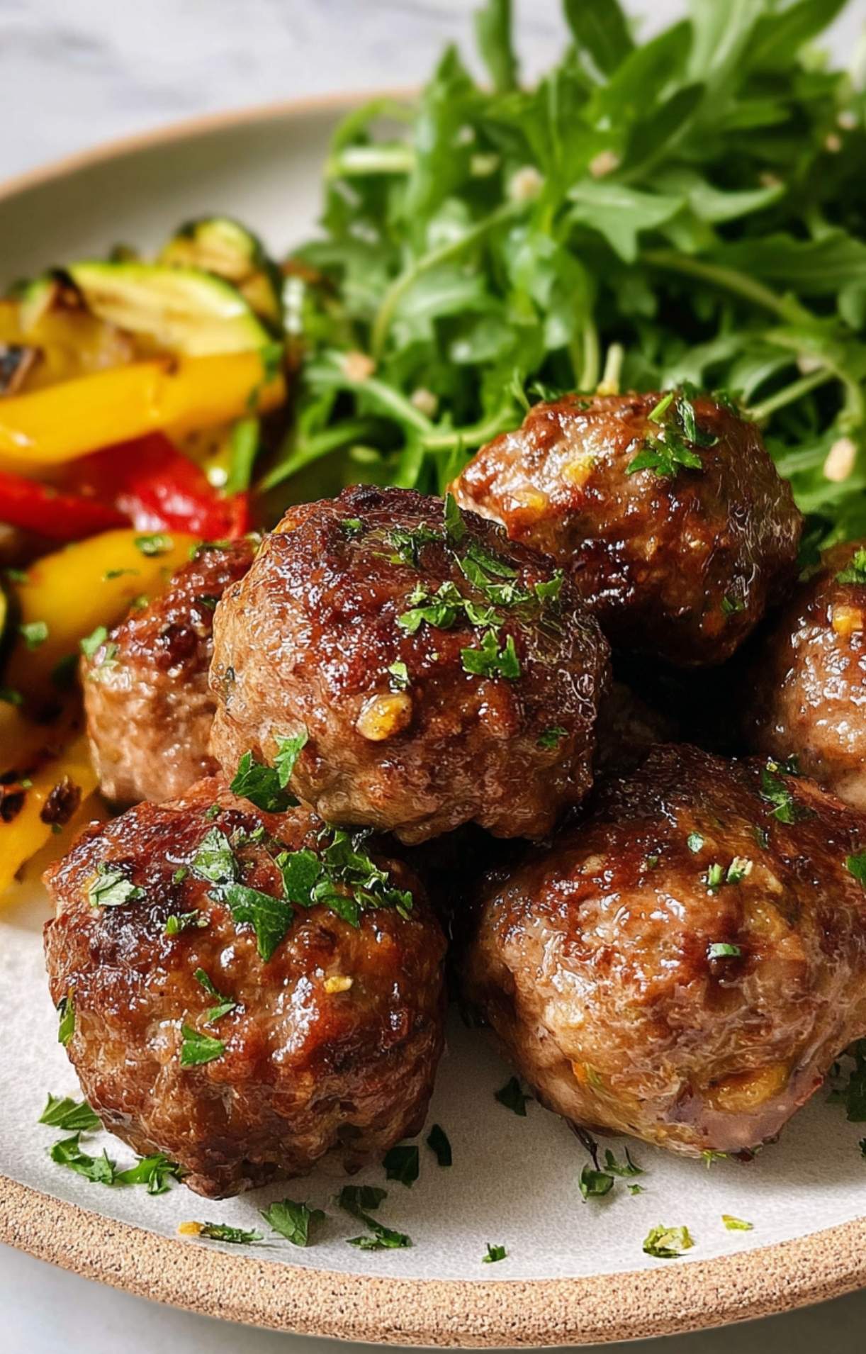 A stack of seasoned air-fried pork meatballs garnished with herbs, served alongside grilled zucchini, peppers, and greens.