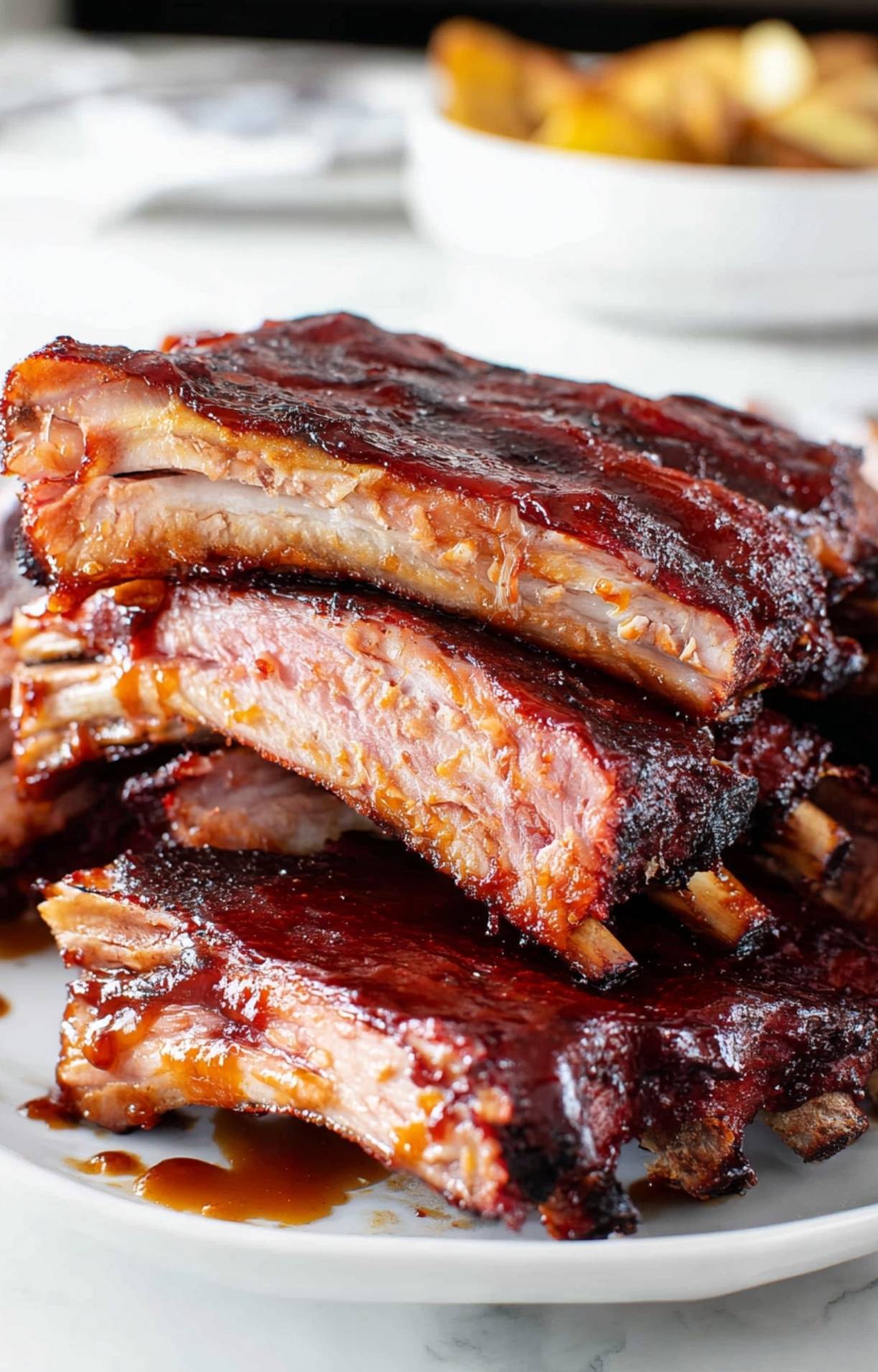 A vertical stack of tender pork ribs served on a white plate with roasted potatoes in the background for a Pellet Smoker Ribs Recipe.