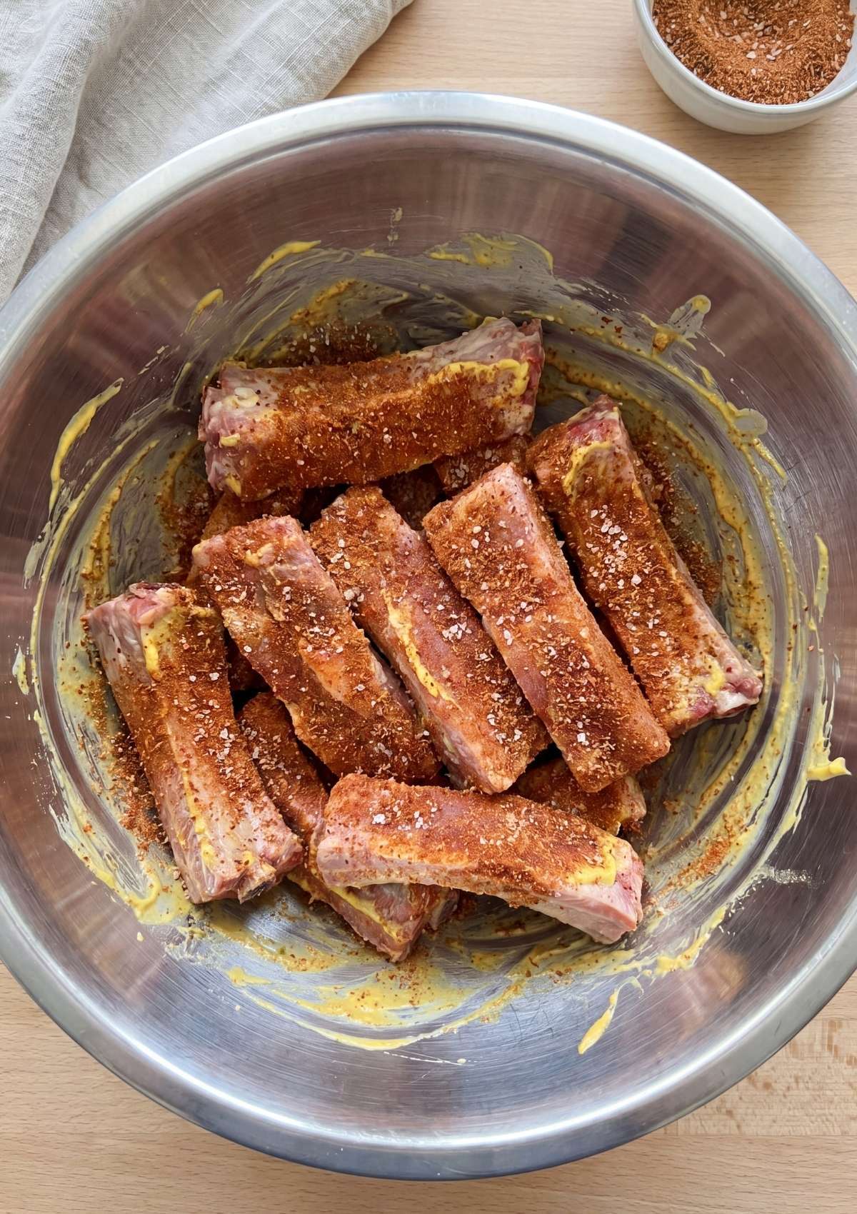 Individual rib segments tossed in a metal bowl with mustard binder and a heavy coating of dry rub and salt.