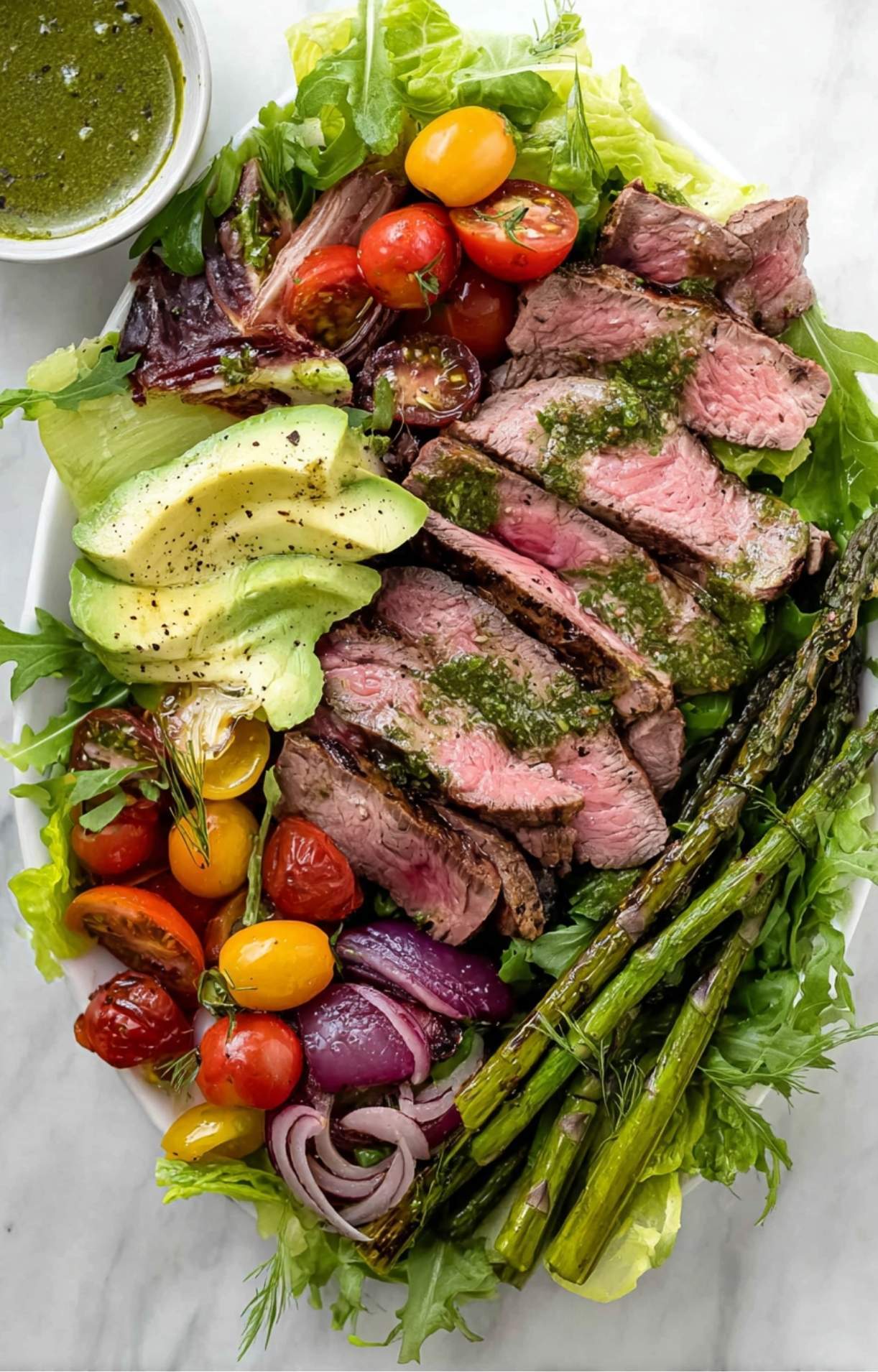 A completed salad platter with sliced steak, grilled asparagus, tomatoes, and avocado, all drizzled with a herb-flecked green dressing.