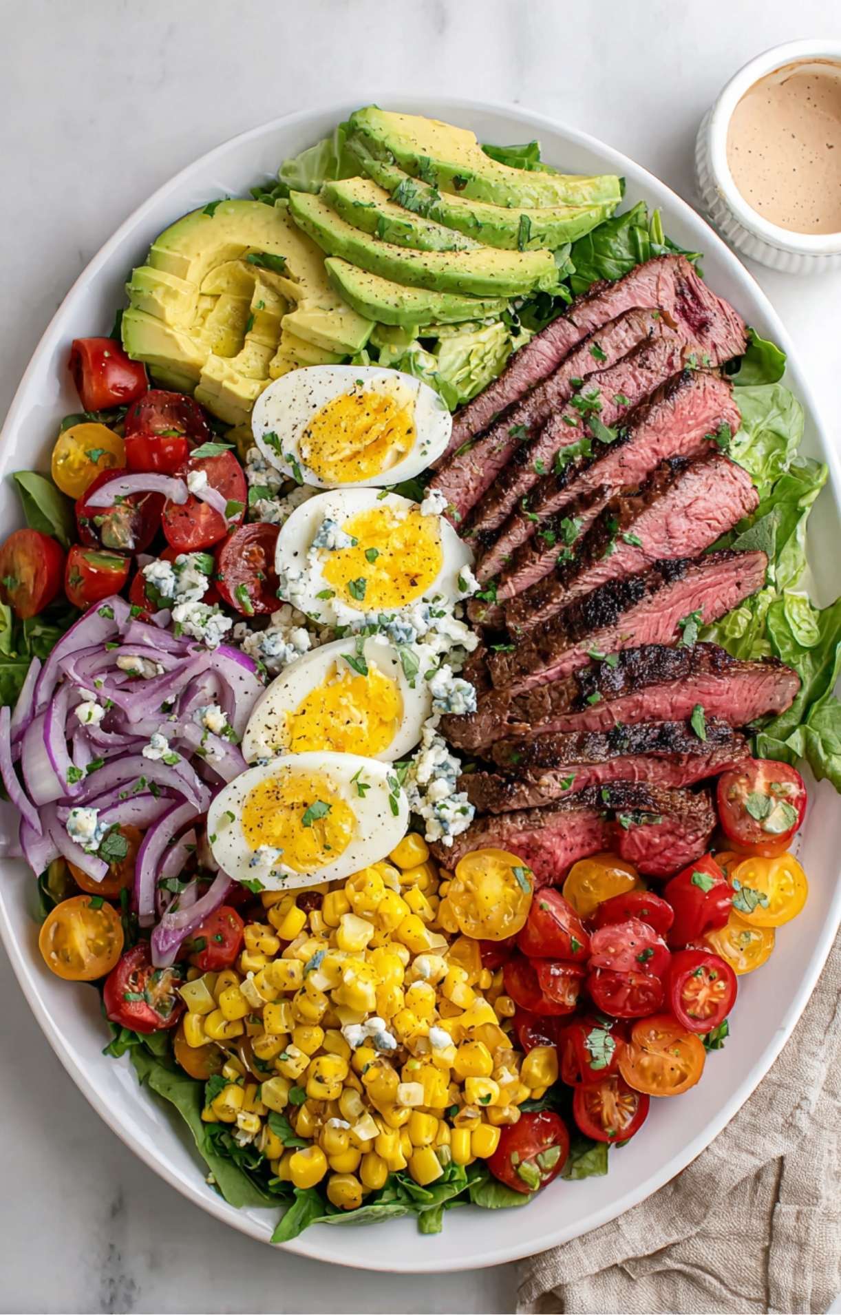 A large oval serving platter beautifully arranged with rows of sliced steak, avocado wedges, boiled eggs, blue cheese, grilled corn, red onions, and tomatoes.