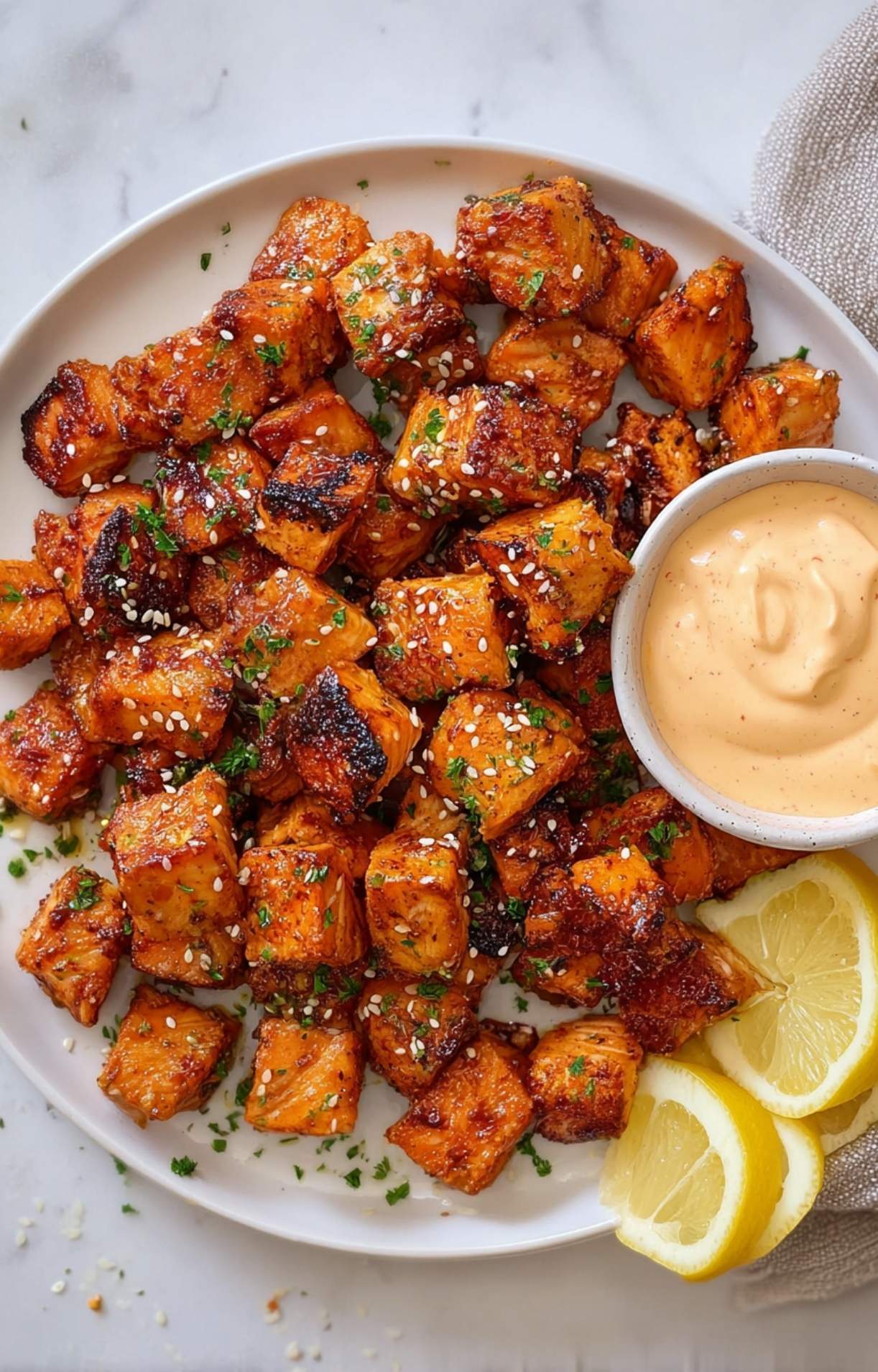 A beautiful presentation of Air Fryer Salmon Bites on a white platter, sprinkled with sesame seeds and parsley, served with lemon slices and a dipping sauce.
